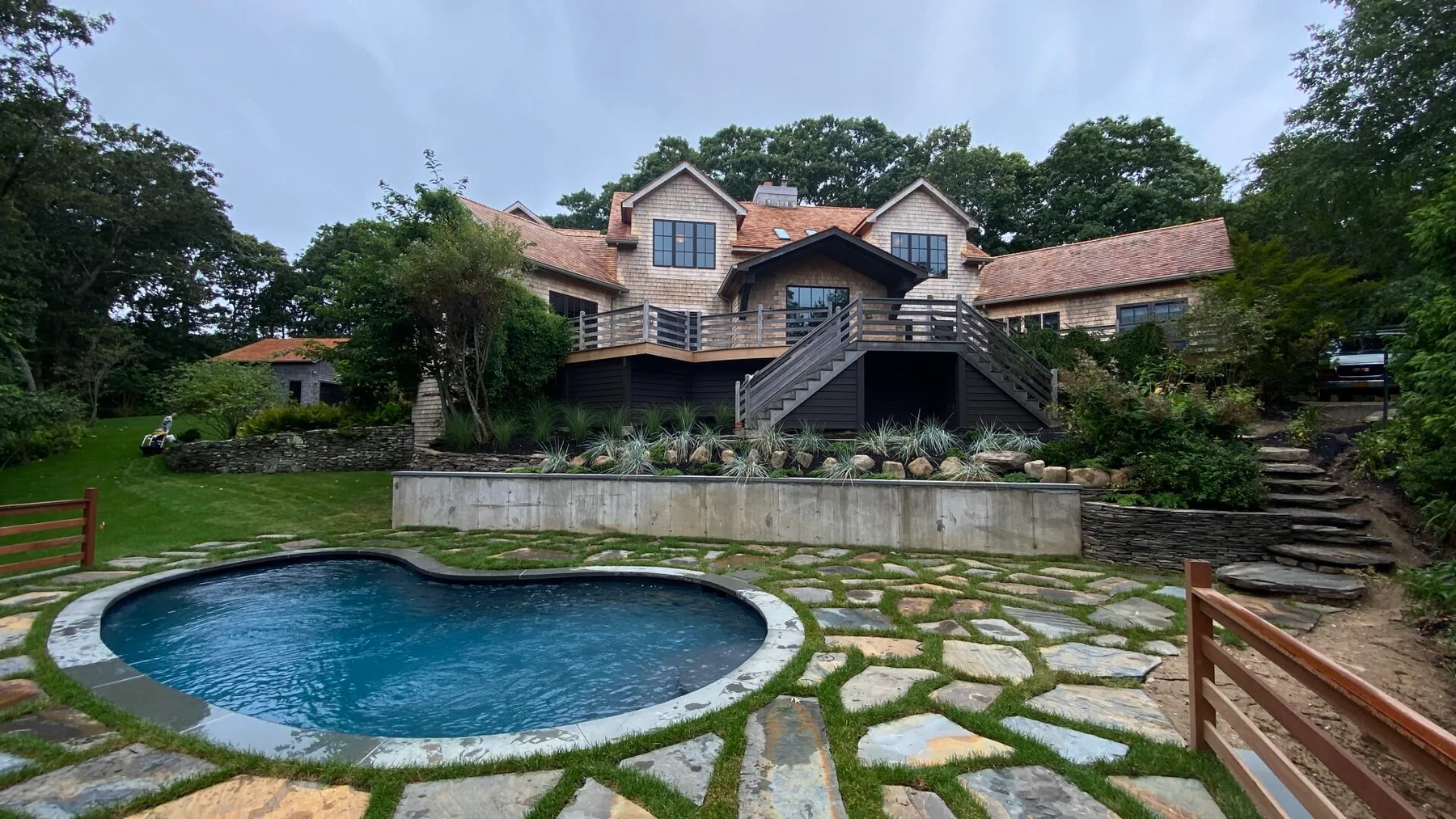 Una piscina al aire libre ajardinada frente a una casa grande y una terraza elevada.