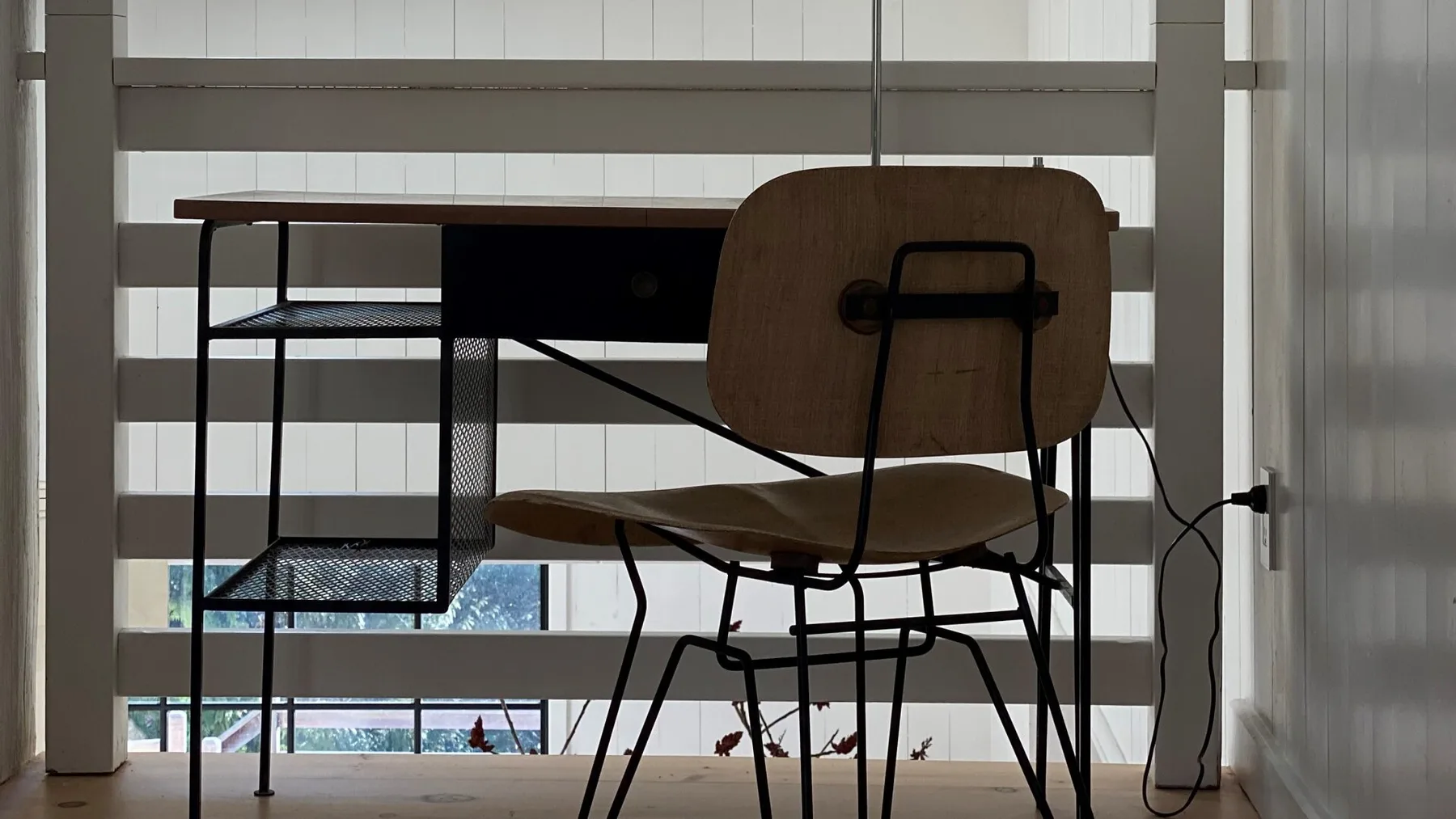 A desk and chair sit in front of large windows with horizontal blinds.