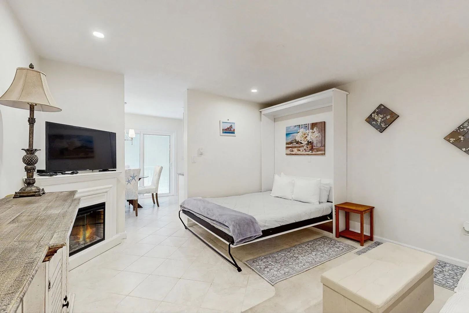 Un dormitorio luminoso con una cama, un televisor y una zona de comedor visible por la puerta.