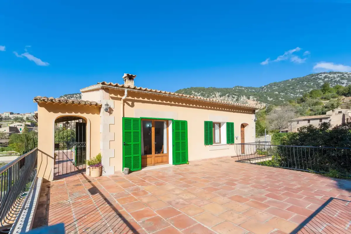 A sunlit house opens onto a broad tiled terrace with green shutters.