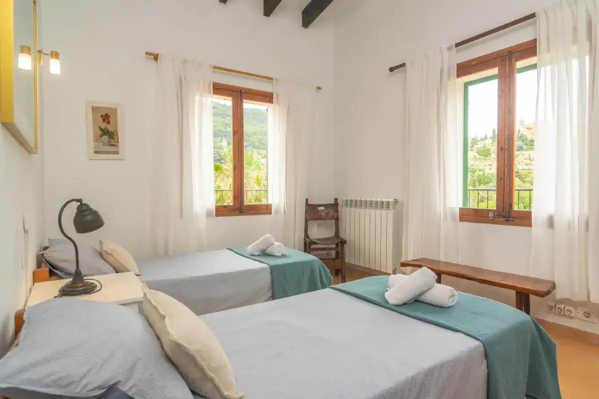 A bright bedroom with two beds and windows overlooking greenery.