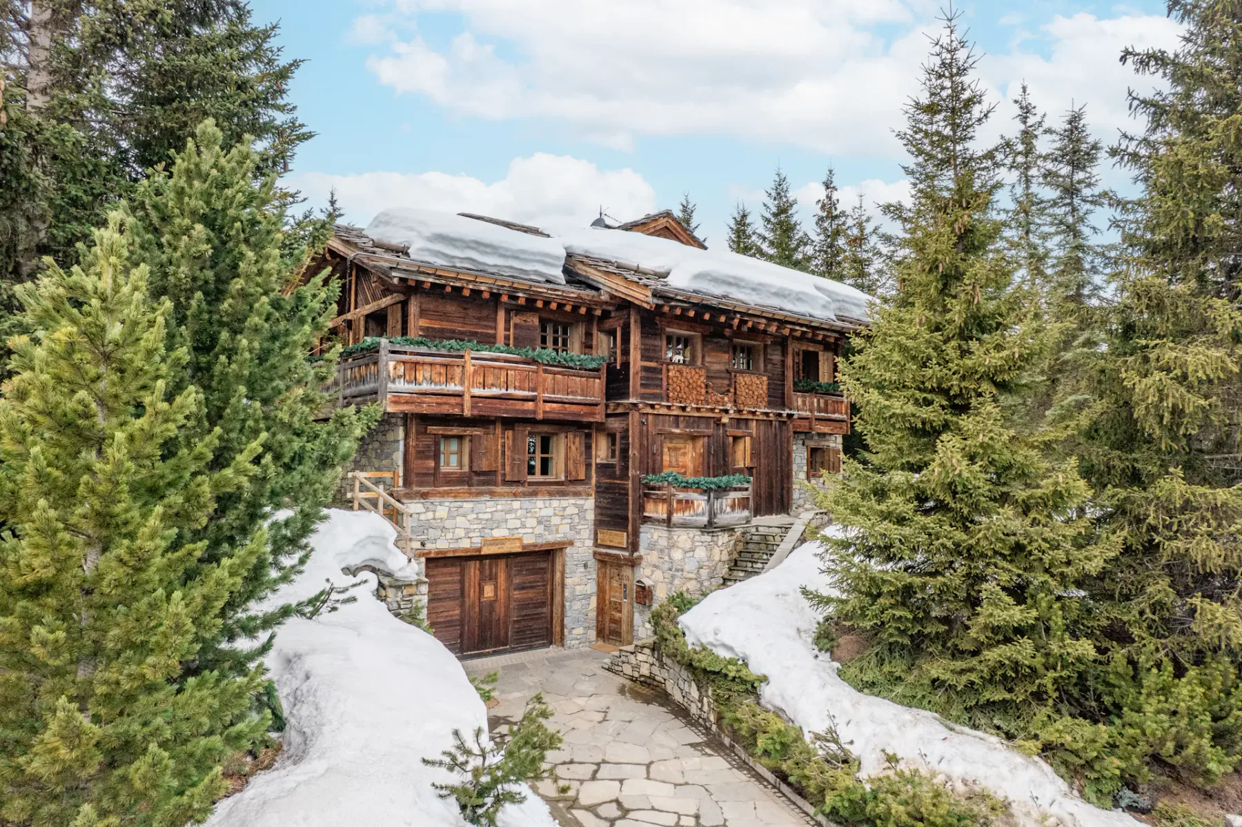A rustic wooden chalet with snow-covered roof, surrounded by evergreen trees and patches of snow, under a partly cloudy sky.