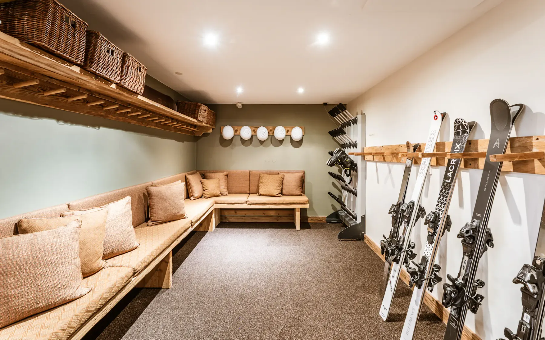 A cozy ski storage room with wooden benches, cushions, wall-mounted ski racks, and wicker baskets on shelves under soft ceiling lights.