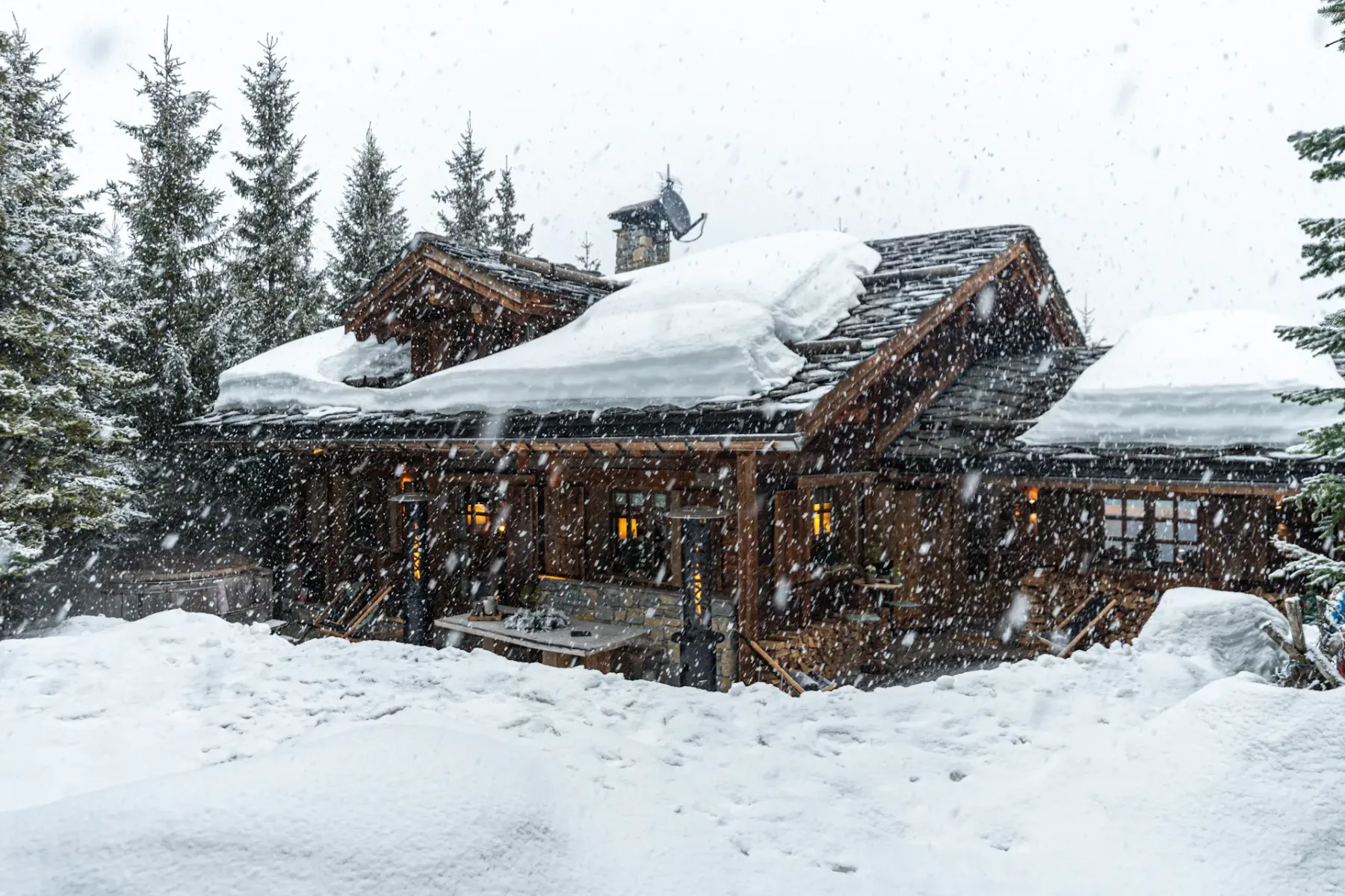 A cozy wooden cabin surrounded by snow-covered trees, with snow falling heavily and a thick layer of snow on the roof.
