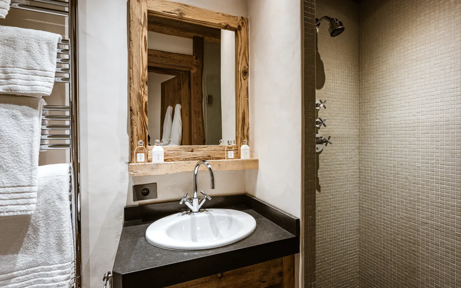 Rustic bathroom with a wooden-framed mirror, white sink, and towels on a rack. A tiled shower area is partially visible.