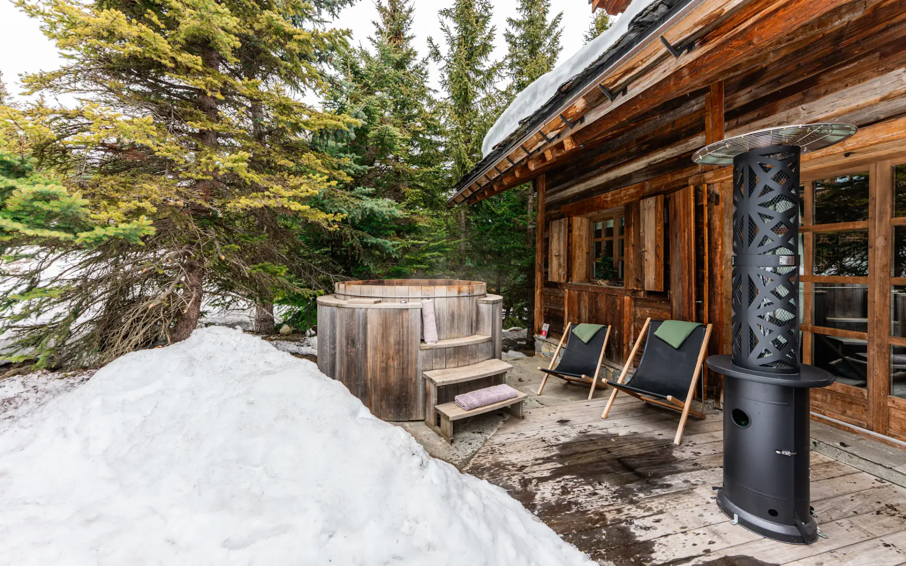 A cozy wooden cabin porch with a hot tub, chairs, and a heater, surrounded by snow and evergreen trees.