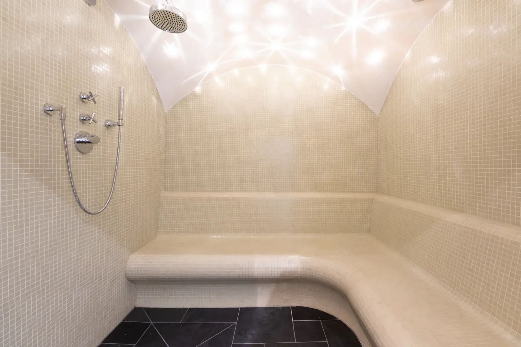 A modern steam room with beige tiled walls and benches, a curved ceiling with star-like lights, and a showerhead on the left wall.