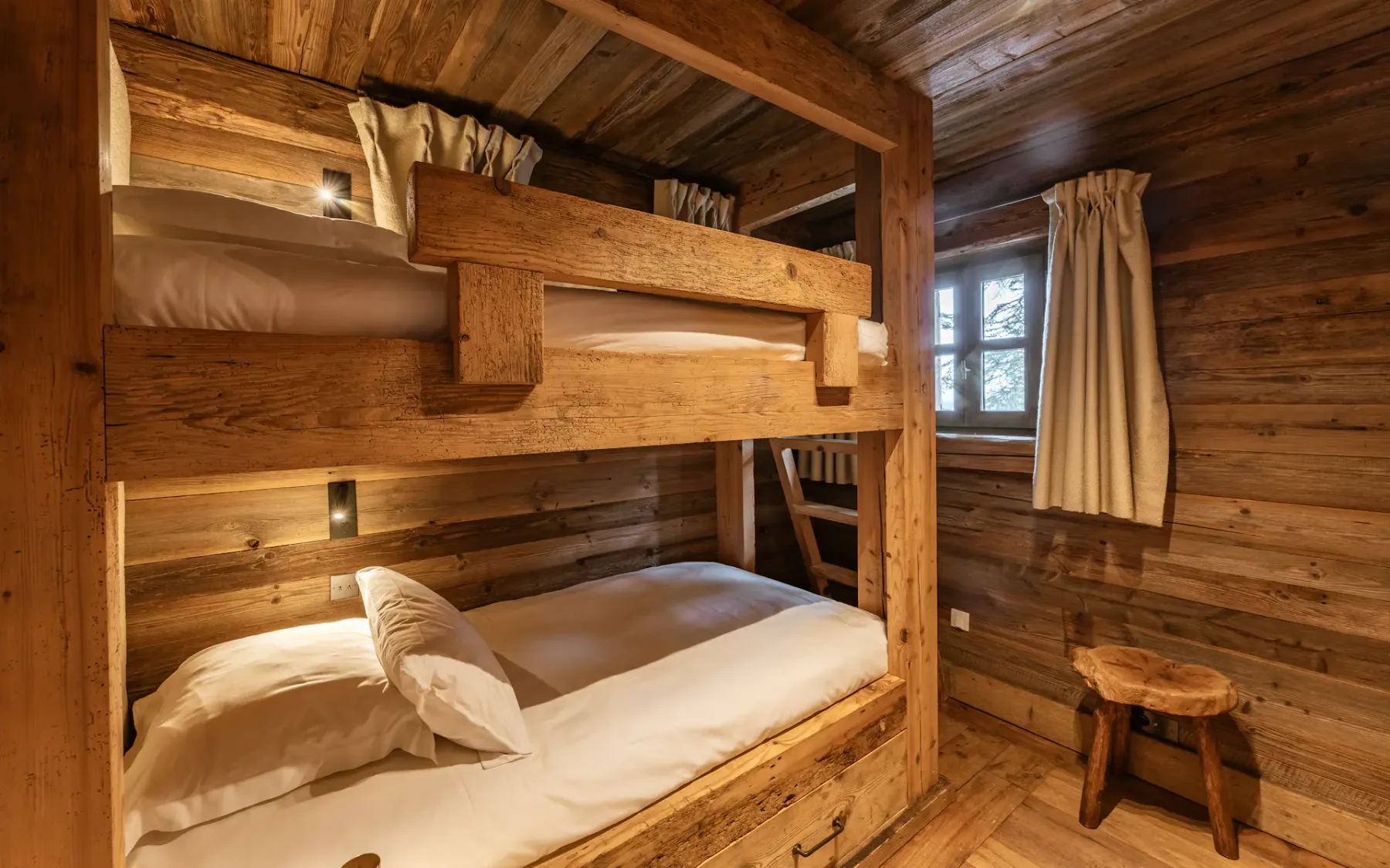 Cozy wooden cabin room with bunk beds, white bedding, rustic decor, small window with curtains, and a wooden stool.