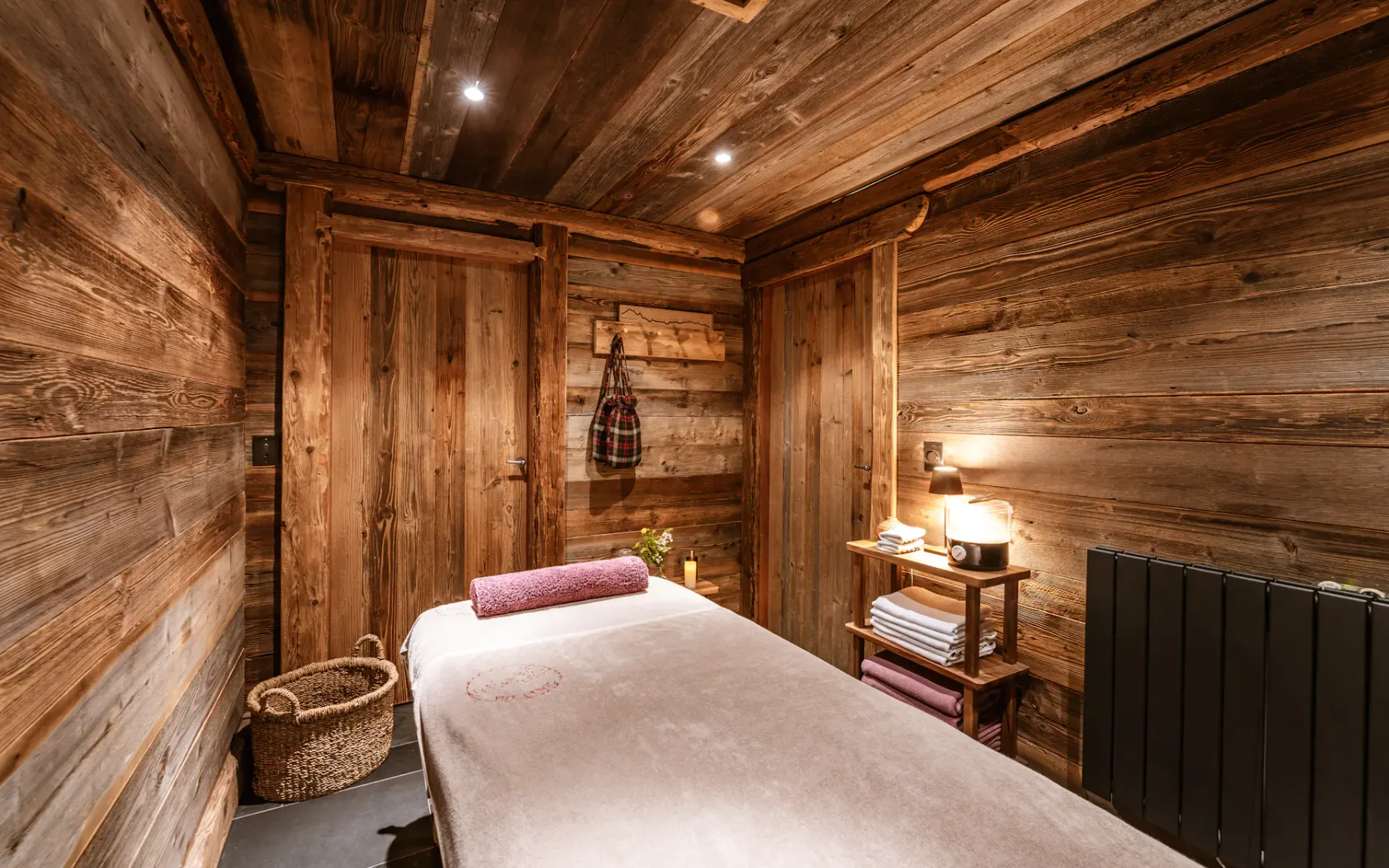 Cozy wooden massage room with a massage table, soft lighting, a basket, and neatly stacked towels on a shelf.