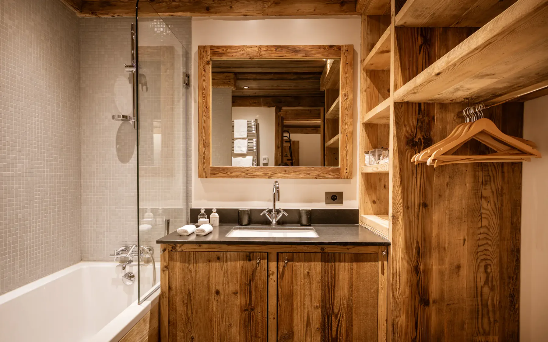 Rustic bathroom with wooden accents, a rectangular mirror, a sink, and a bathtub with a glass screen. Open shelves hold towels and hangers.
