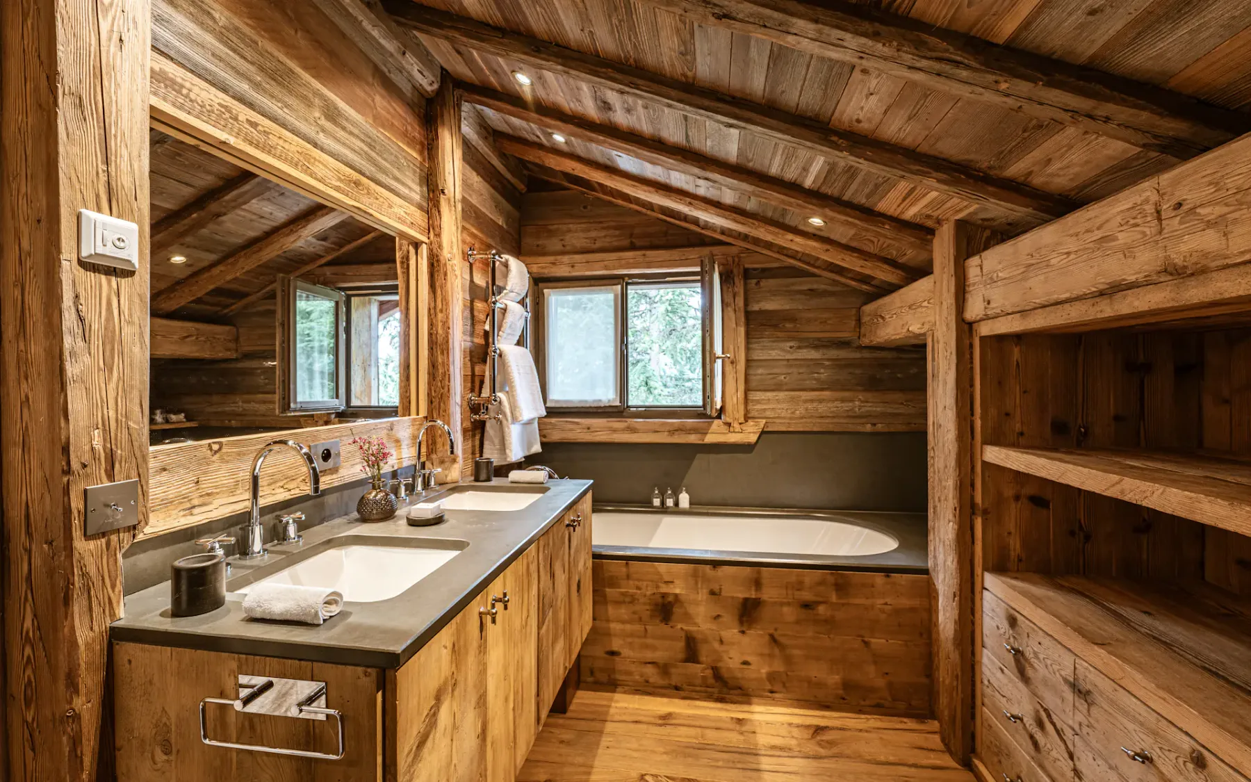 Rustic wooden bathroom with double sinks, a bathtub, and open shelves. Sloped ceiling and a window provide natural light.
