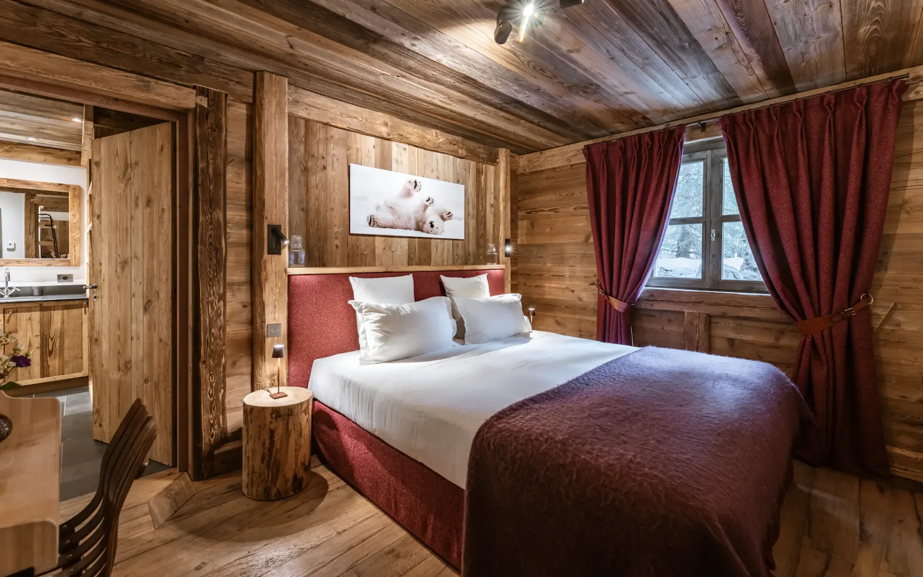 Cozy rustic bedroom with wooden walls, a large bed with red accents, a window with red curtains, and a small wooden table. Warm lighting enhances the ambiance.