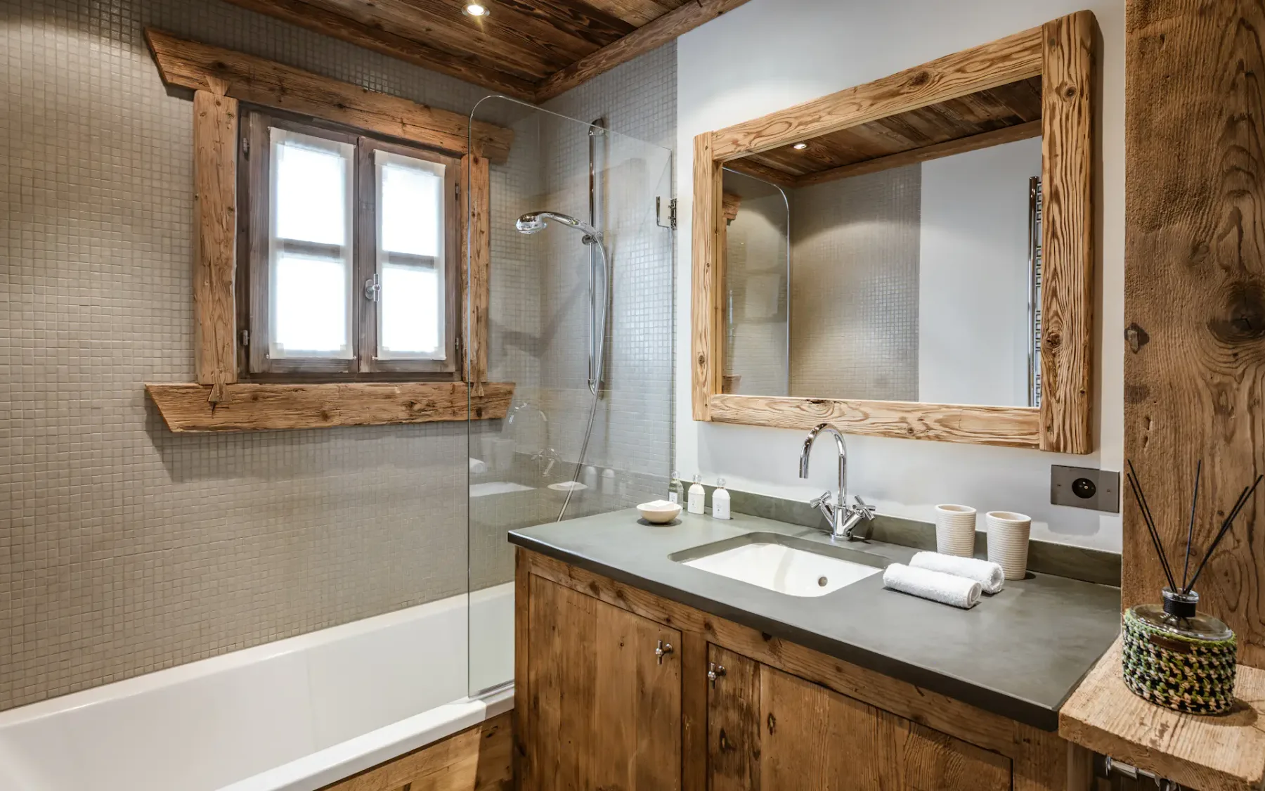 Rustic bathroom with wooden accents, featuring a vanity with a large mirror, a window, and a shower-tub combo with glass partition.