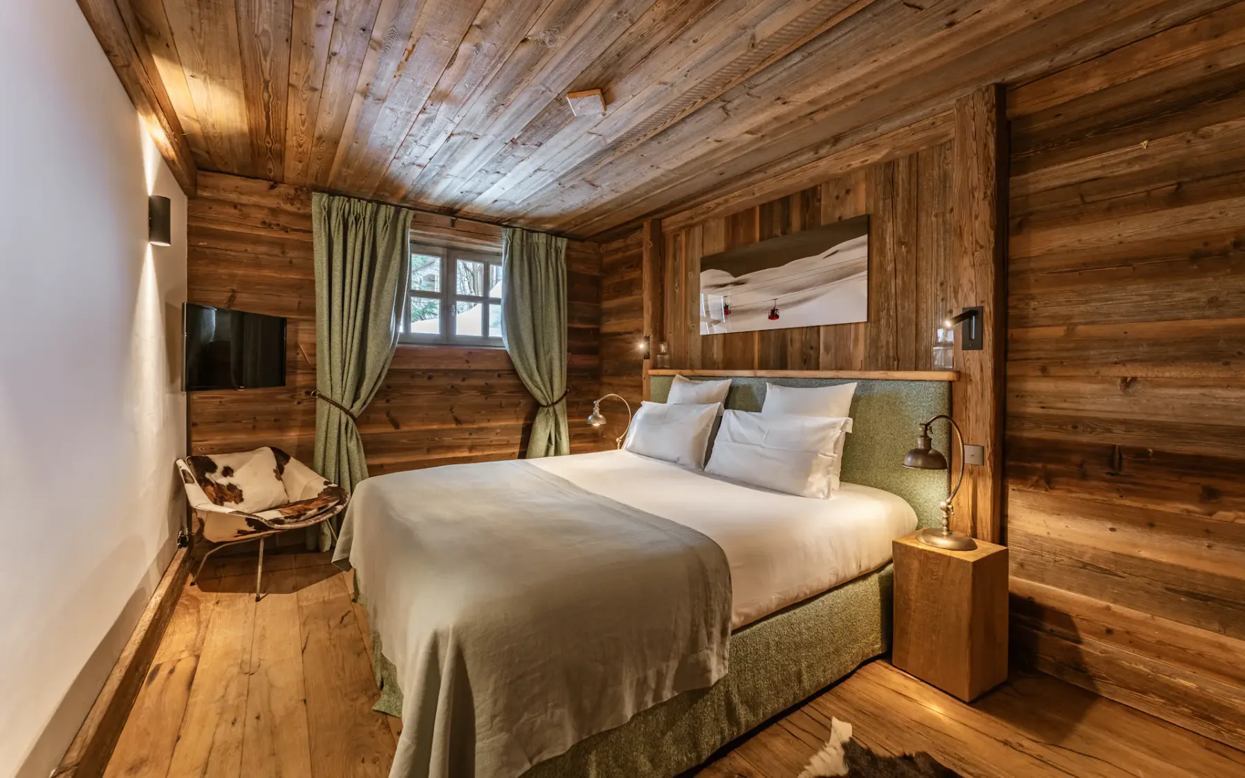 Cozy rustic bedroom with wooden walls and ceiling, a large bed, green curtains, a wall-mounted TV, and a small chair with a fur throw.