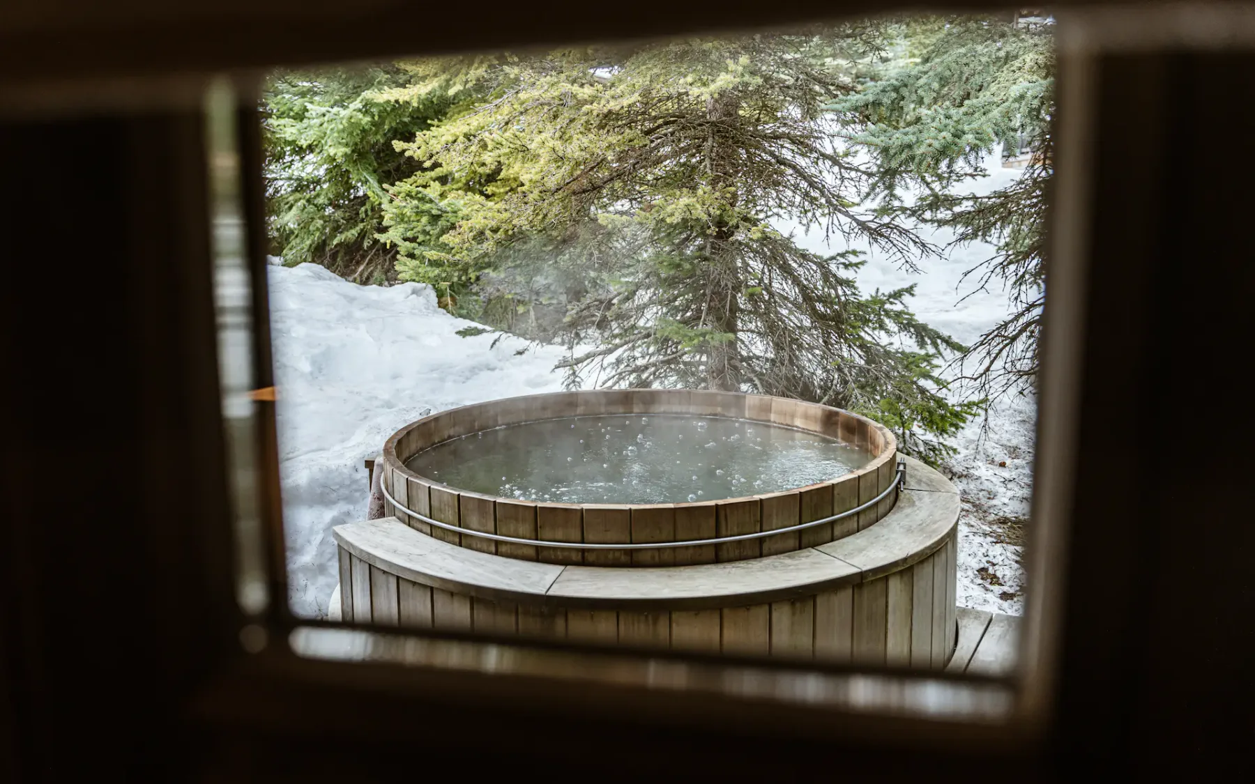 Wooden hot tub steaming outdoors, surrounded by snow and evergreen trees, viewed through a window.
