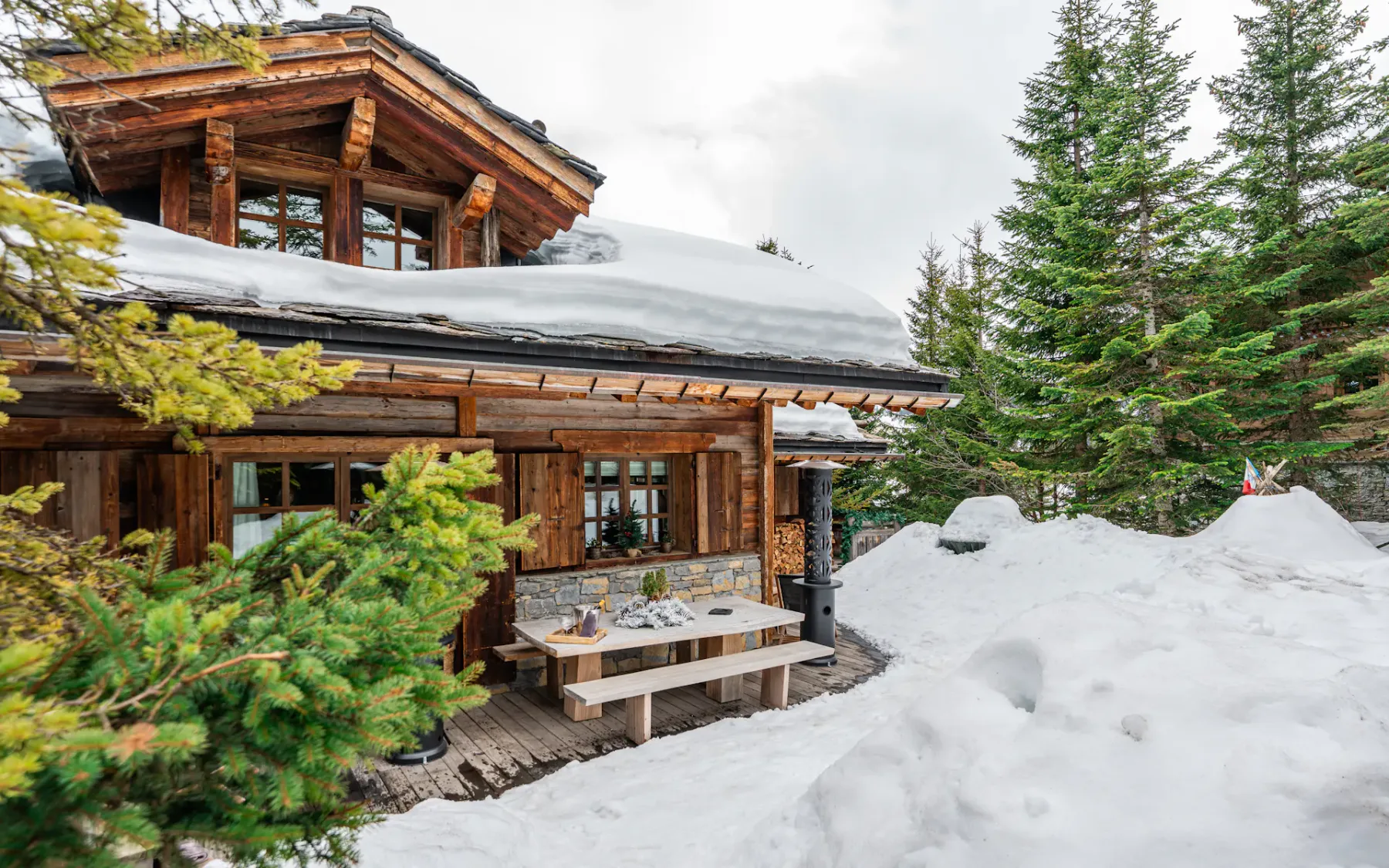 Snow-covered wooden cabin with a porch, surrounded by pine trees and mounds of snow, under a cloudy sky.