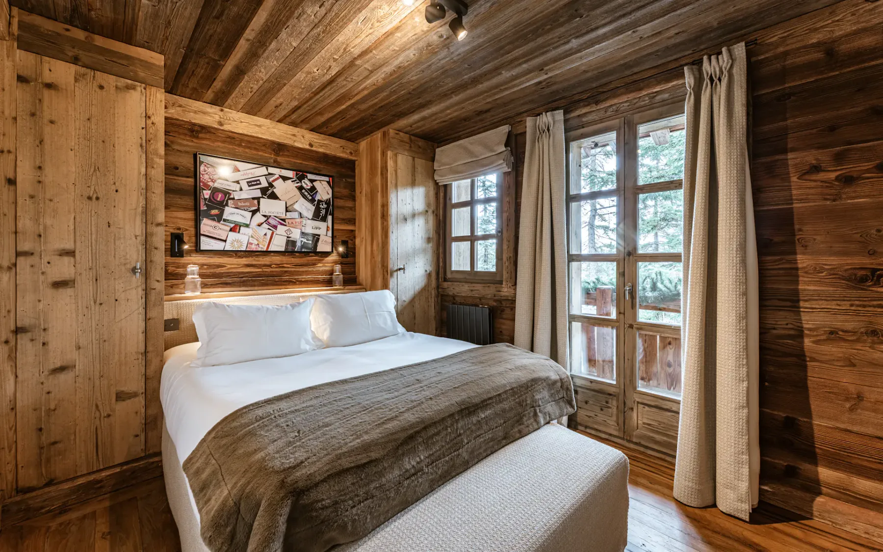 Cozy wooden bedroom with a double bed, white linens, brown throw, large window with curtains, and artwork above the bed.