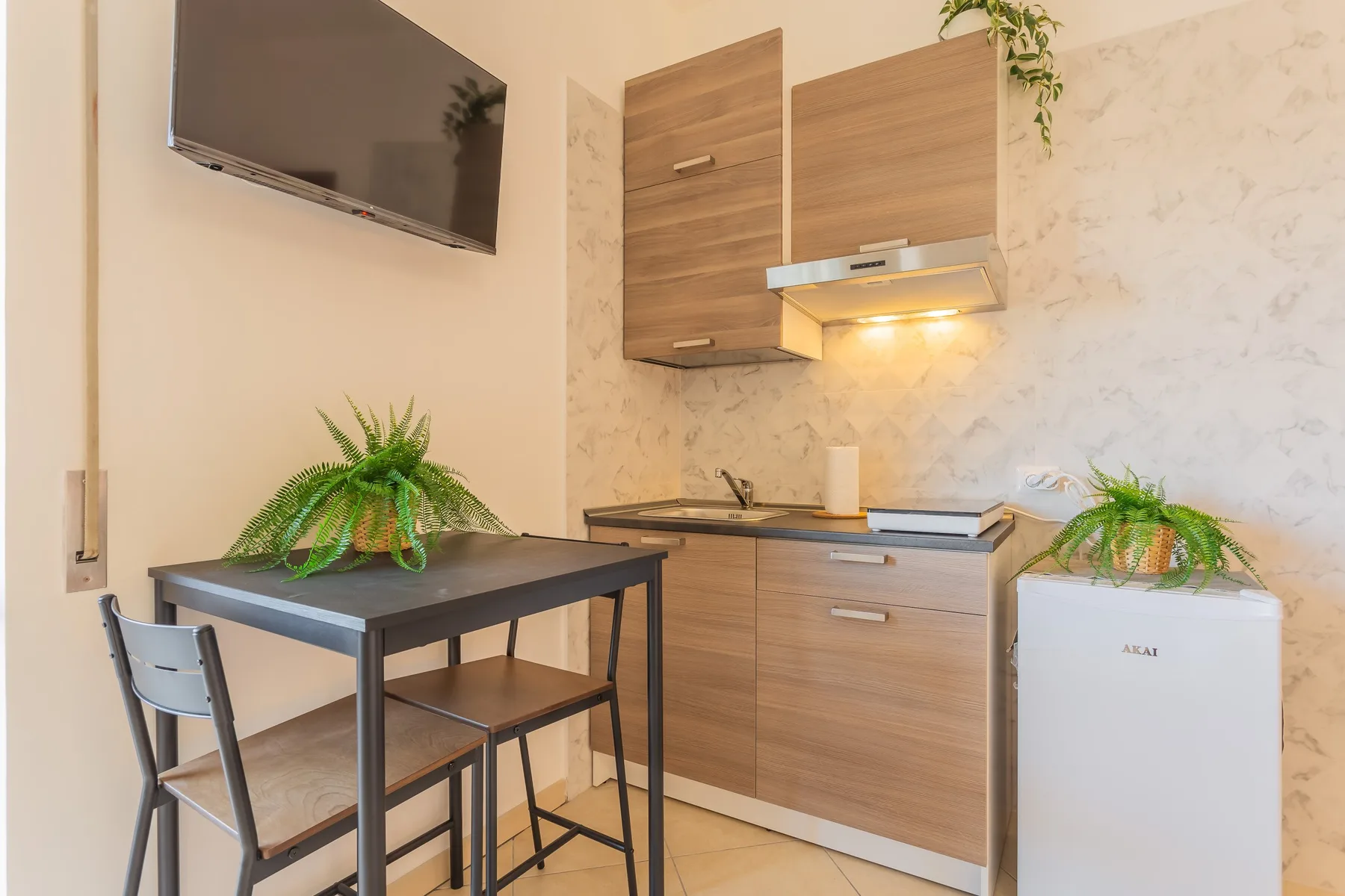 A compact kitchenette with a dining table, wall-mounted television, and potted plants.