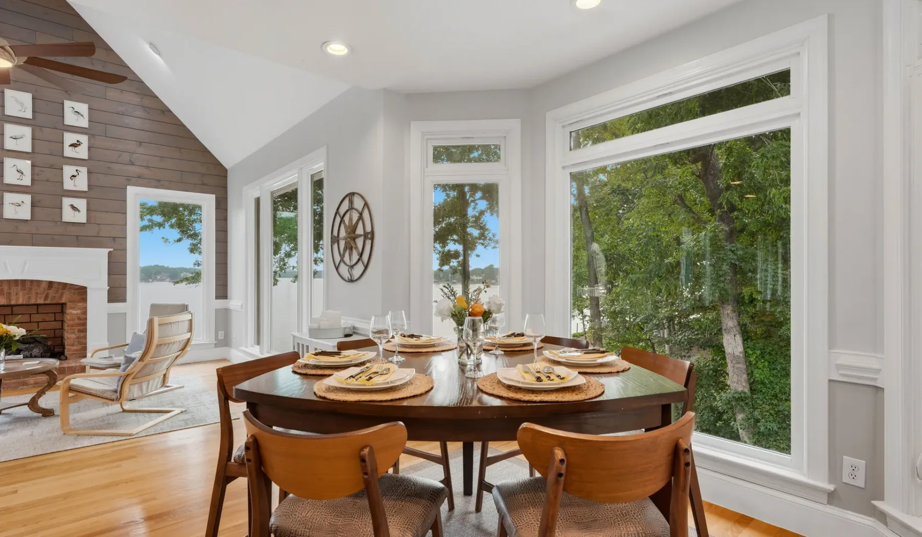 A dining area with a wooden table beside large windows overlooking trees.