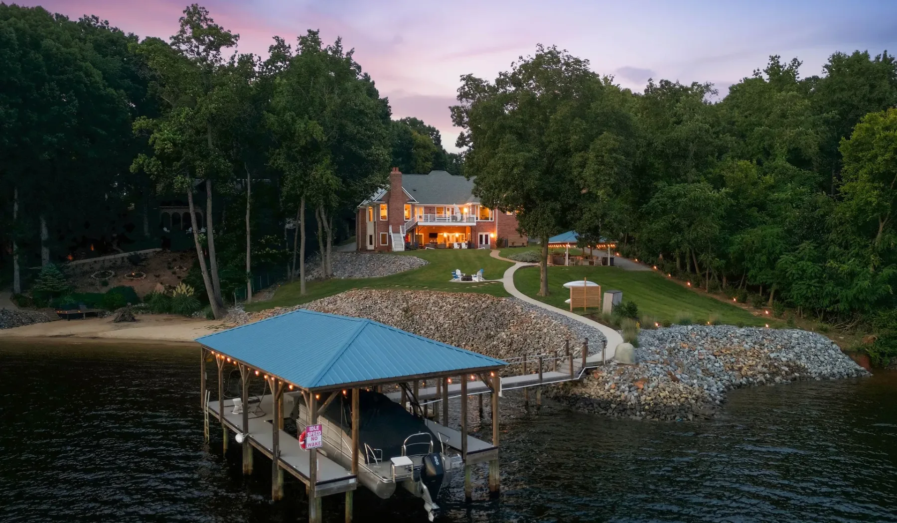 A lakeside house and covered dock sit beside a wooded shoreline at dusk.