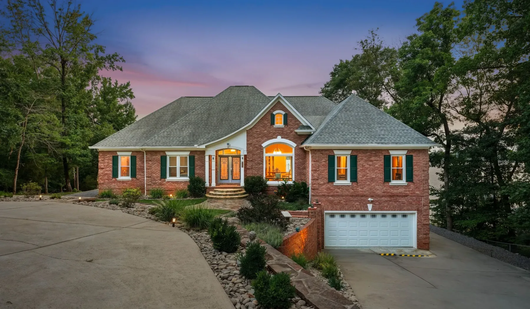 A brick house with a gray roof stands beside a curved driveway at dusk.