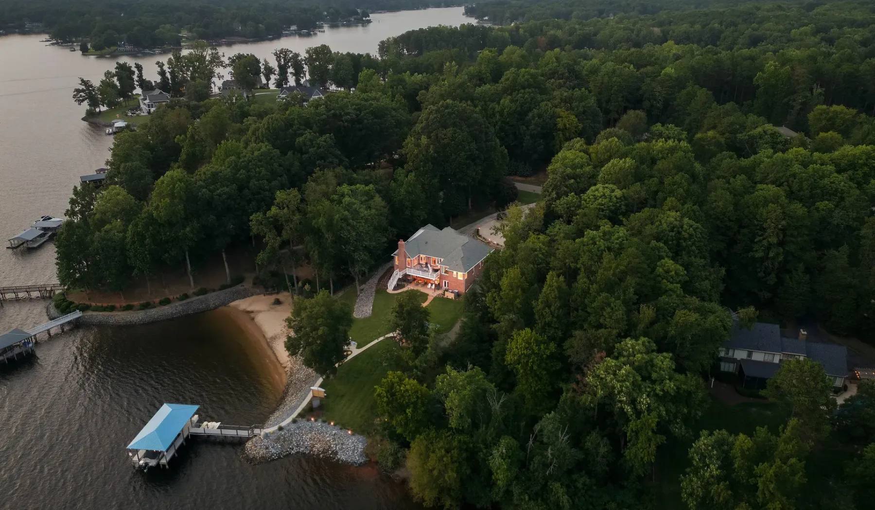 An aerial view shows a house surrounded by trees beside a wooded shoreline.
