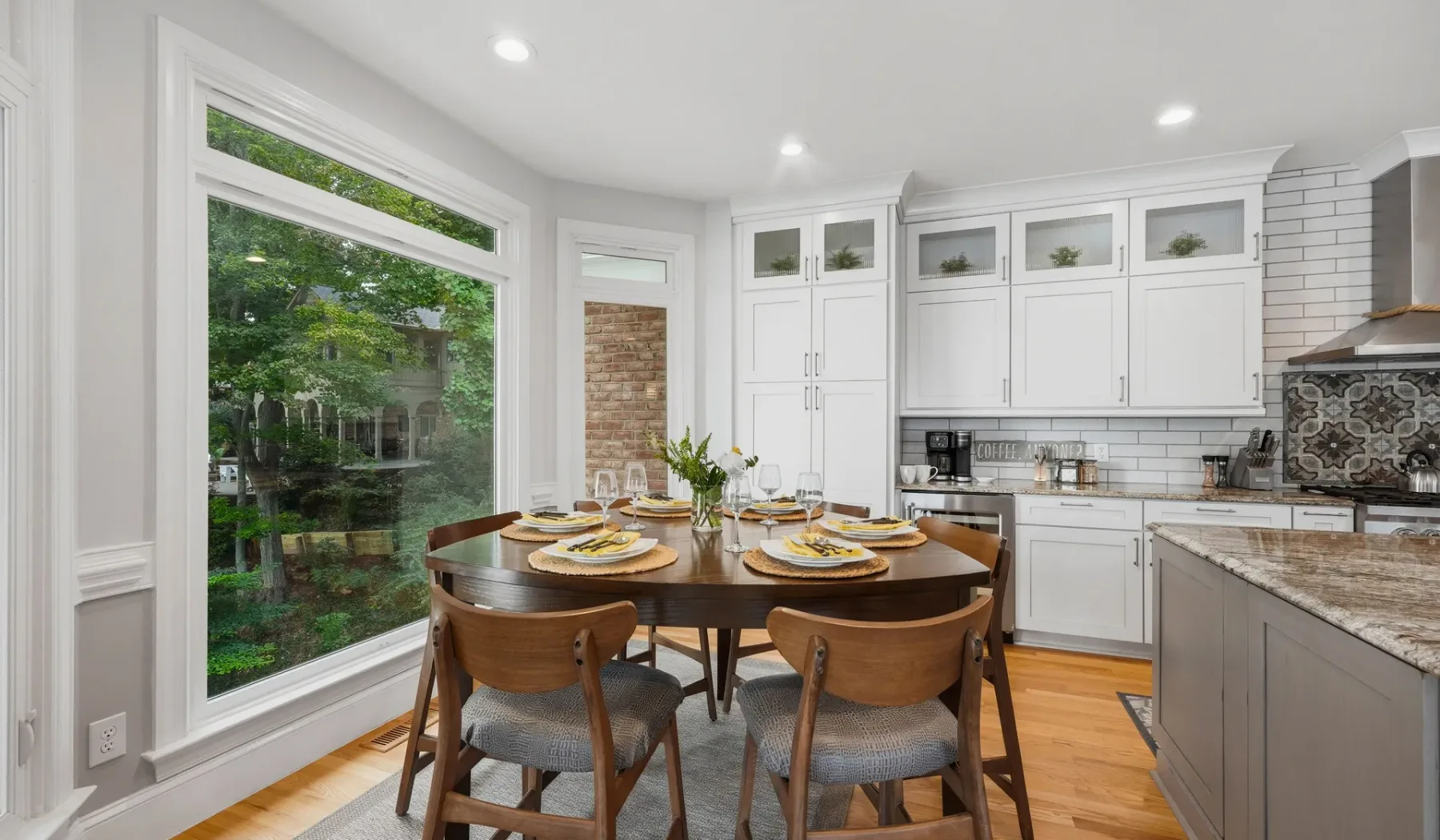 A dining area sits beside a bright kitchen, with large windows overlooking greenery.