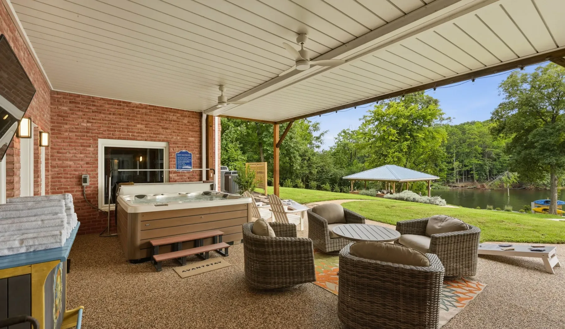 A covered outdoor patio has seating beside a hot tub, with a lawn and pond beyond.