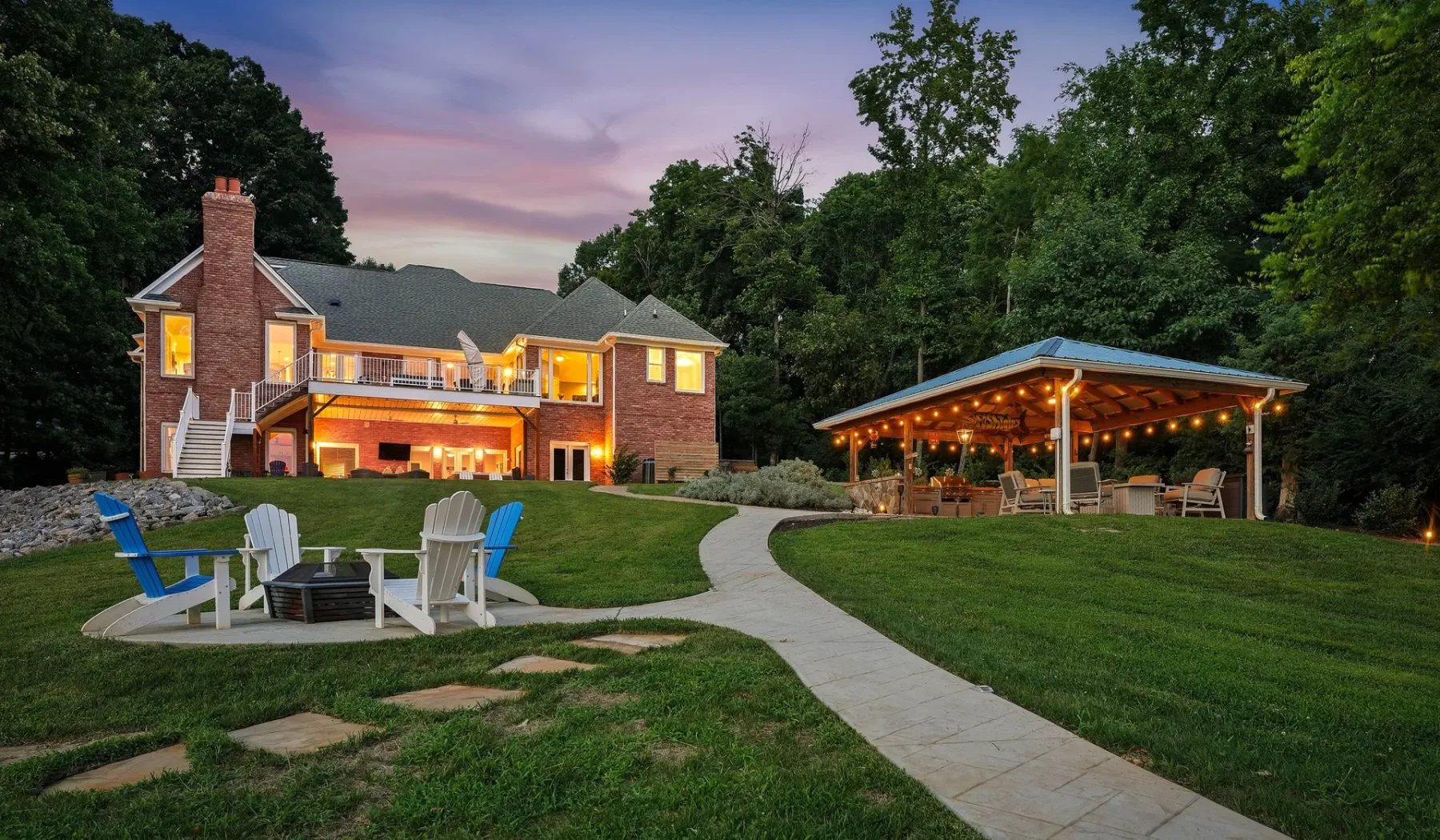 A landscaped outdoor seating area and lit pavilion beside a large house at dusk.