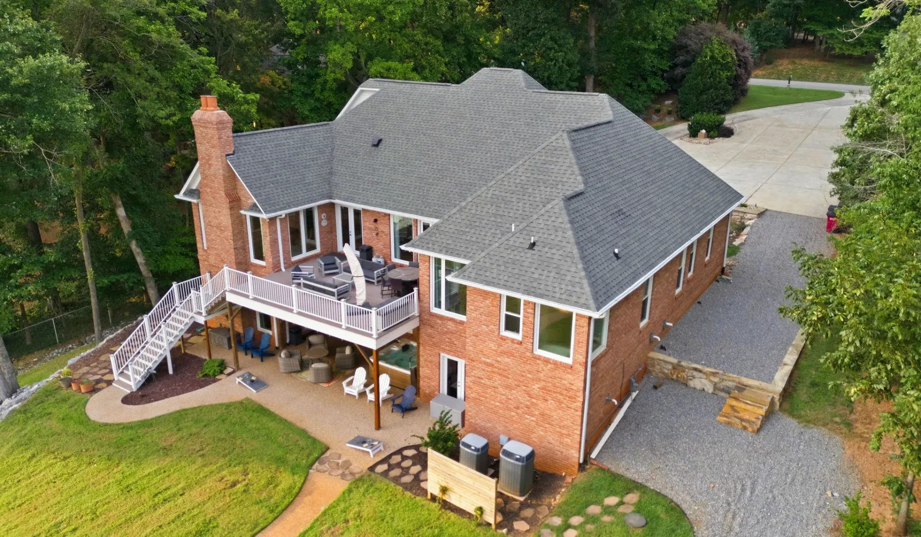Aerial view of a brick house with a large gray roof, deck, and surrounding trees.