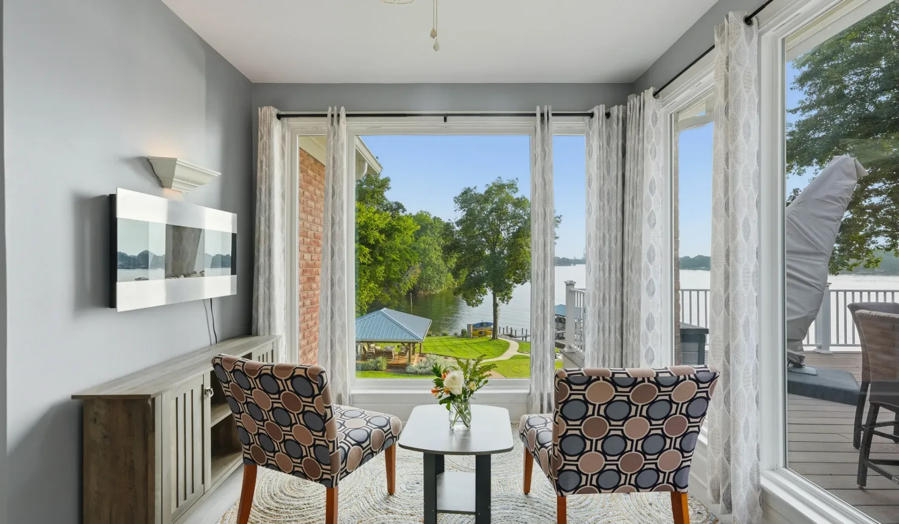 A bright living room with two patterned seats beside large windows overlooking greenery and water.
