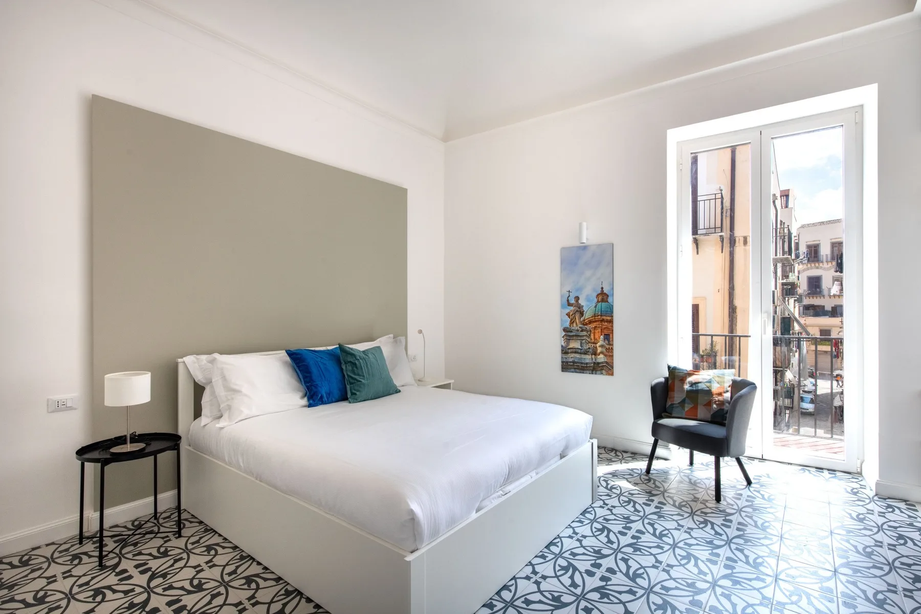 Un dormitorio luminoso con una cama blanca, suelo estampado y puertas altas de cristal.