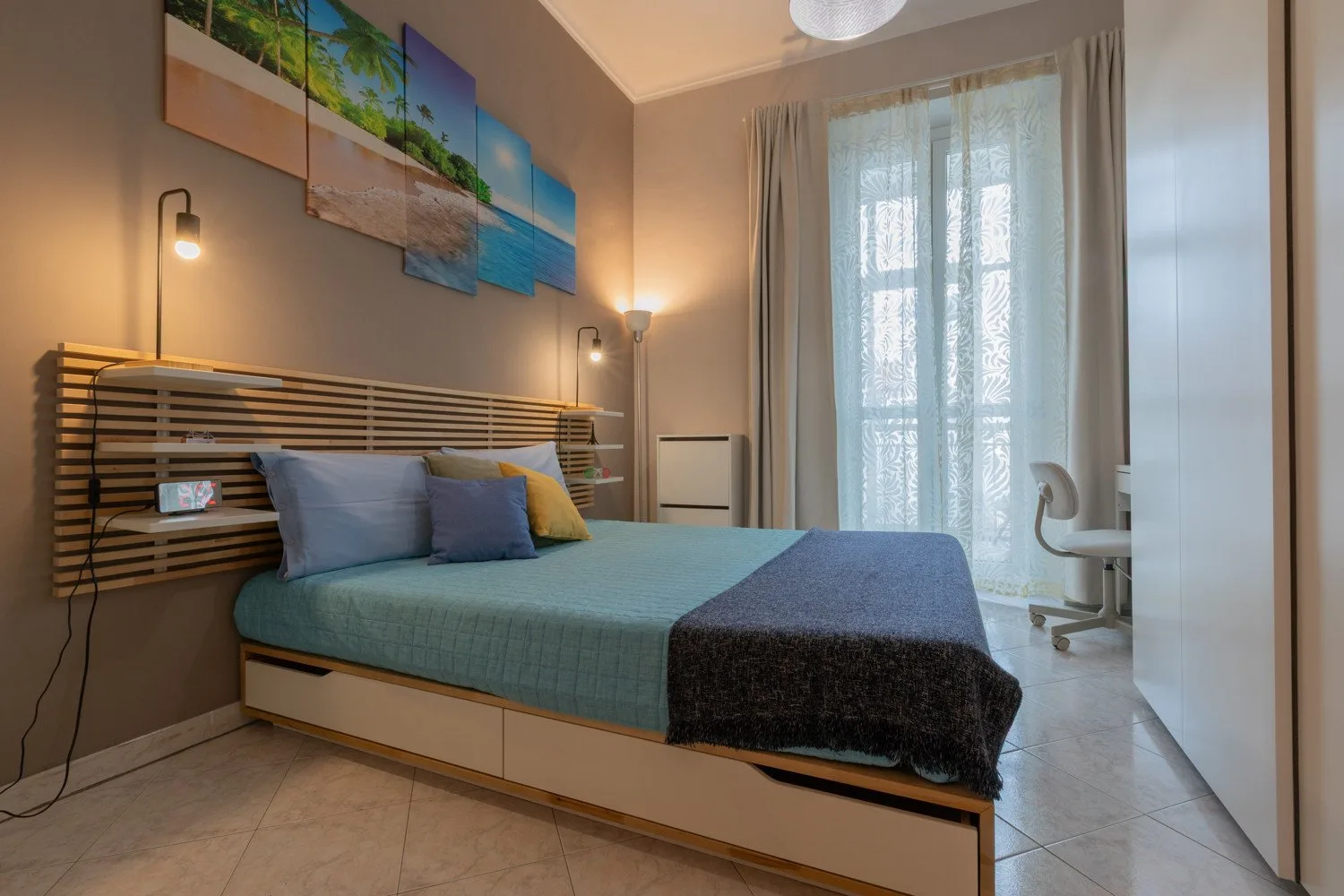 A bedroom with a bed, colorful wall art, and a bright curtained window.