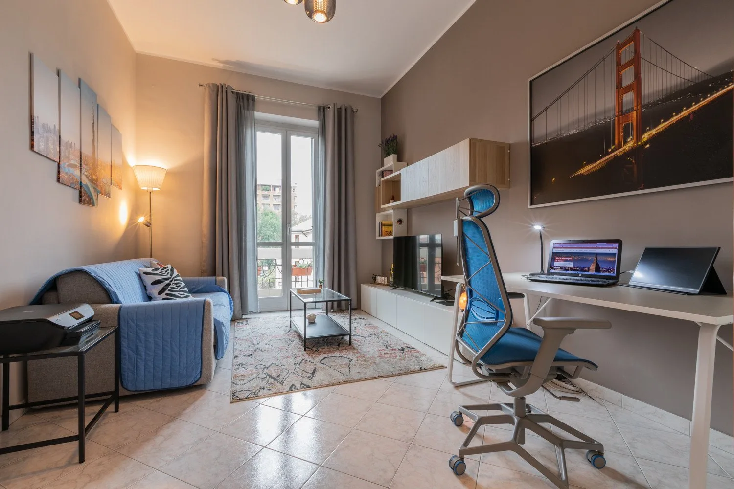 A living room with a blue sofa, television, and desk beside tall windows.