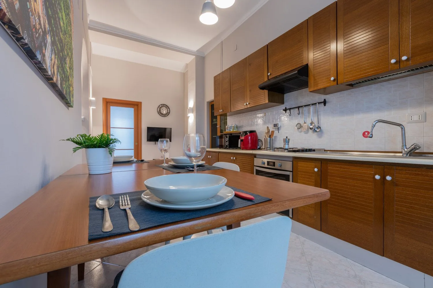 A dining table set with dishes stands beside a kitchen with wooden cabinets.