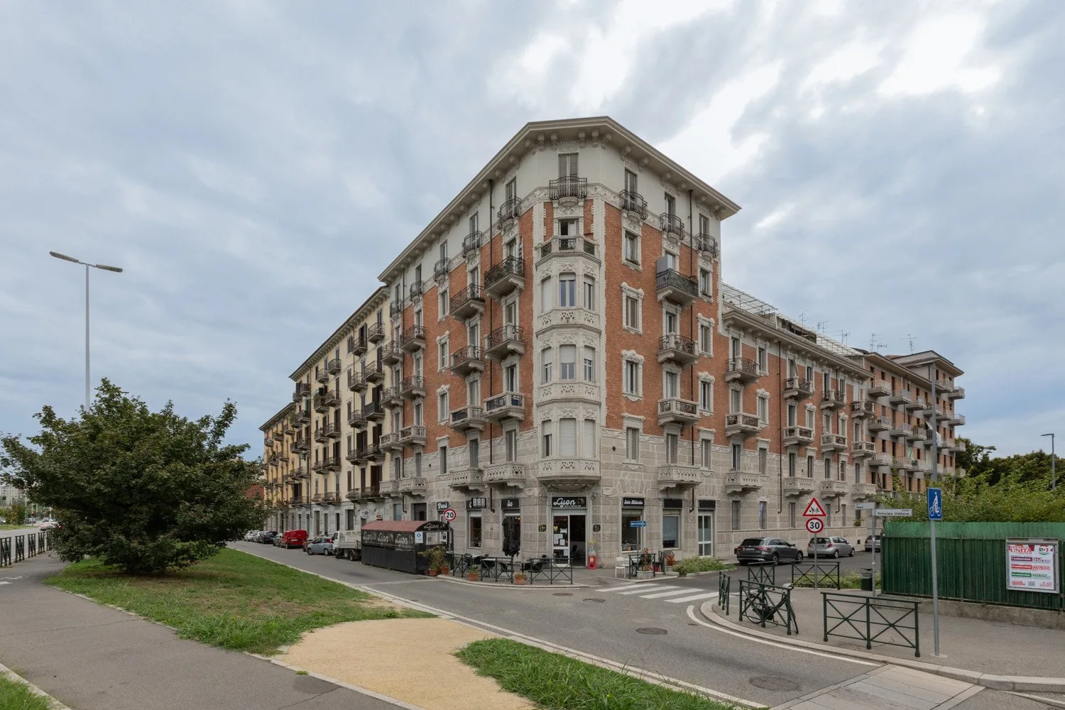 A large corner apartment building stands beside a street under a cloudy sky.