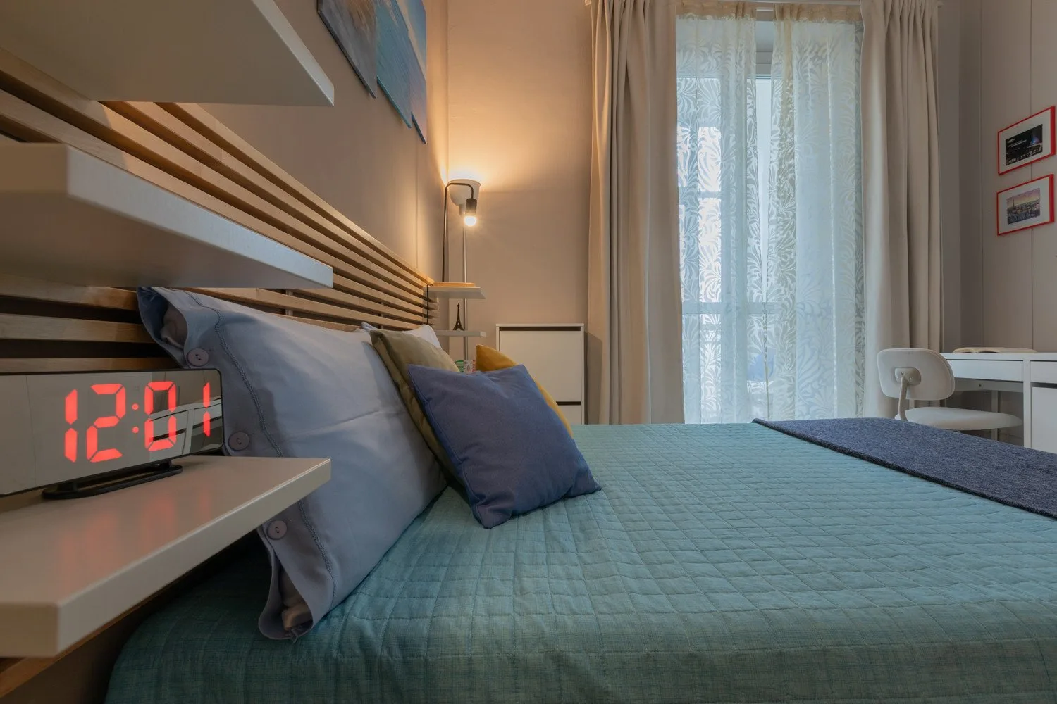A bedroom with a bed covered in teal bedding beside a curtained window.