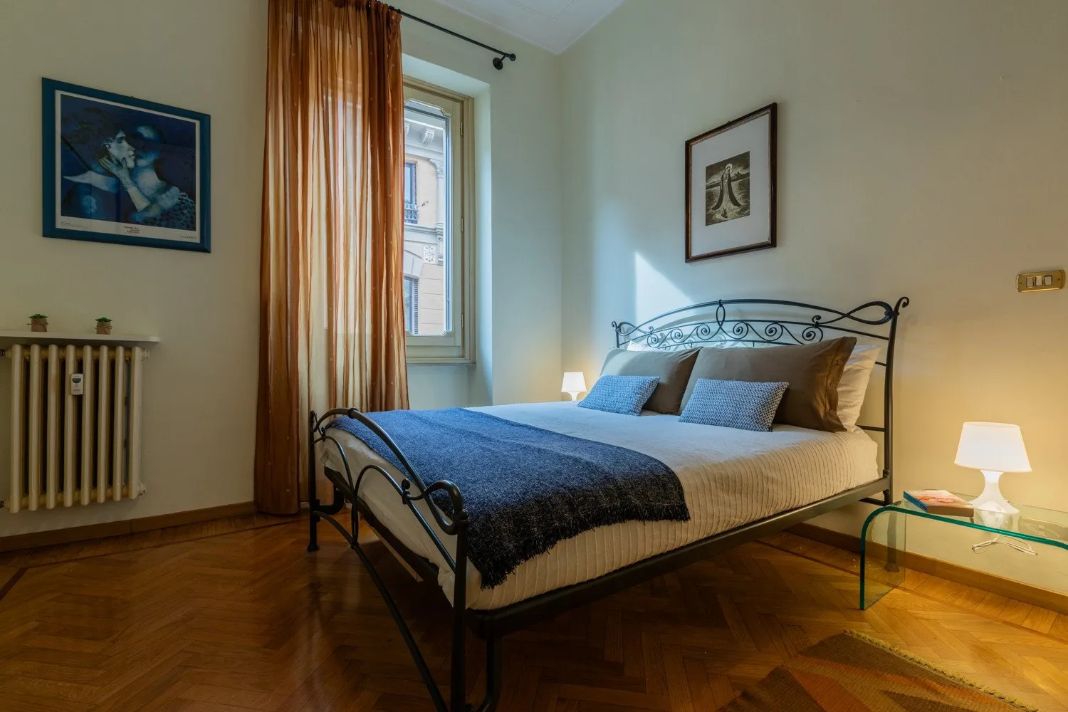 Un dormitorio con una cama de estructura metálica, ropa de cama azul y una ventana con cortinas.