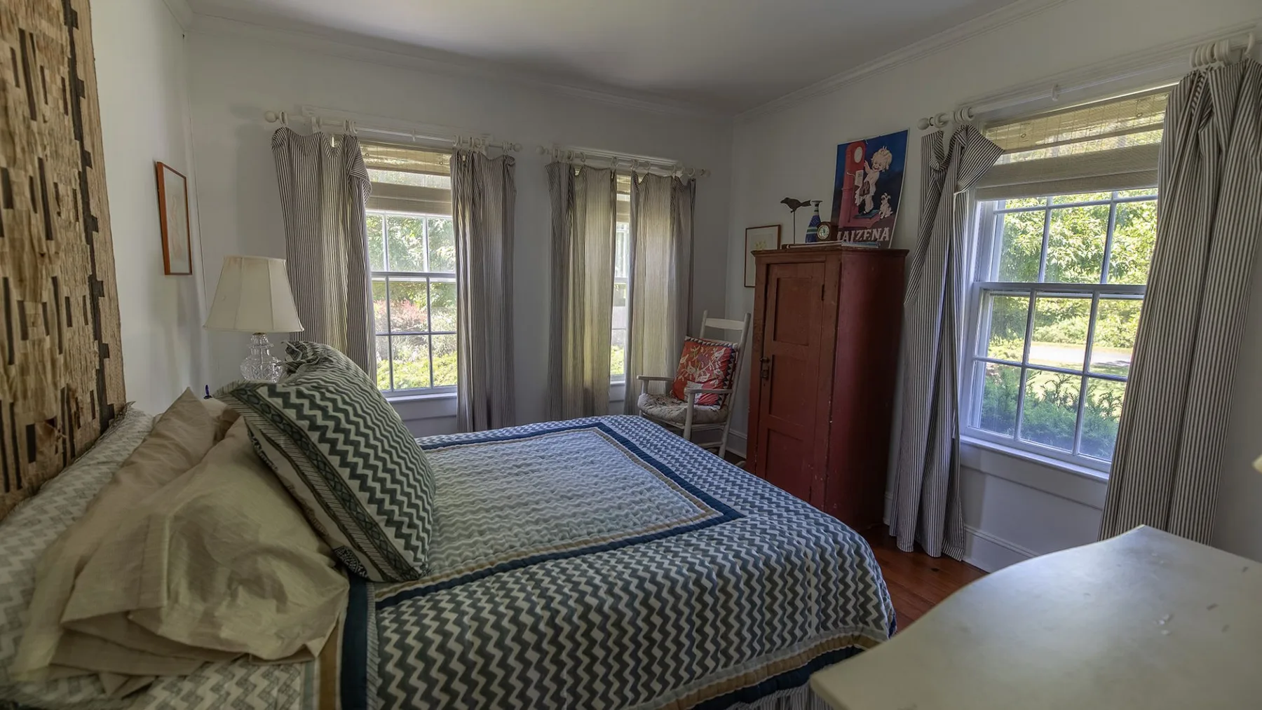 Un dormitorio con una cama estampada, ventanas con cortinas y un armario de madera.