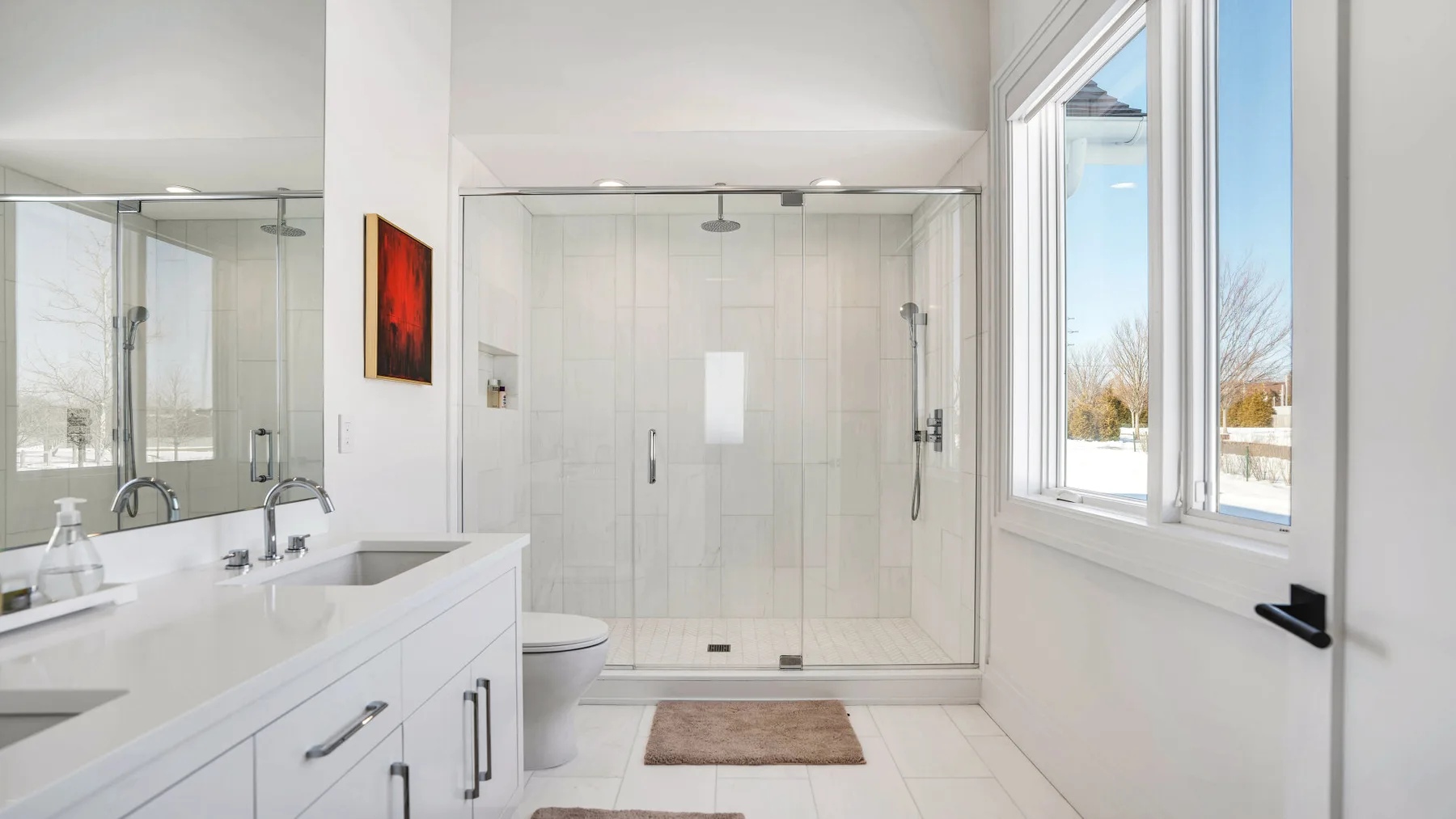 Un baño luminoso con ducha de vidrio, tocador blanco y una ventana grande.