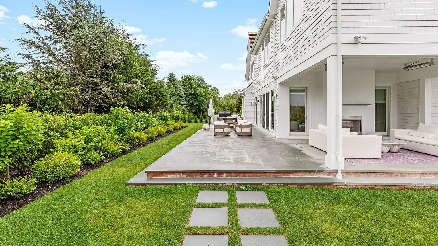 Un patio ajardinado junto a una casa blanca, con asientos al aire libre y césped.