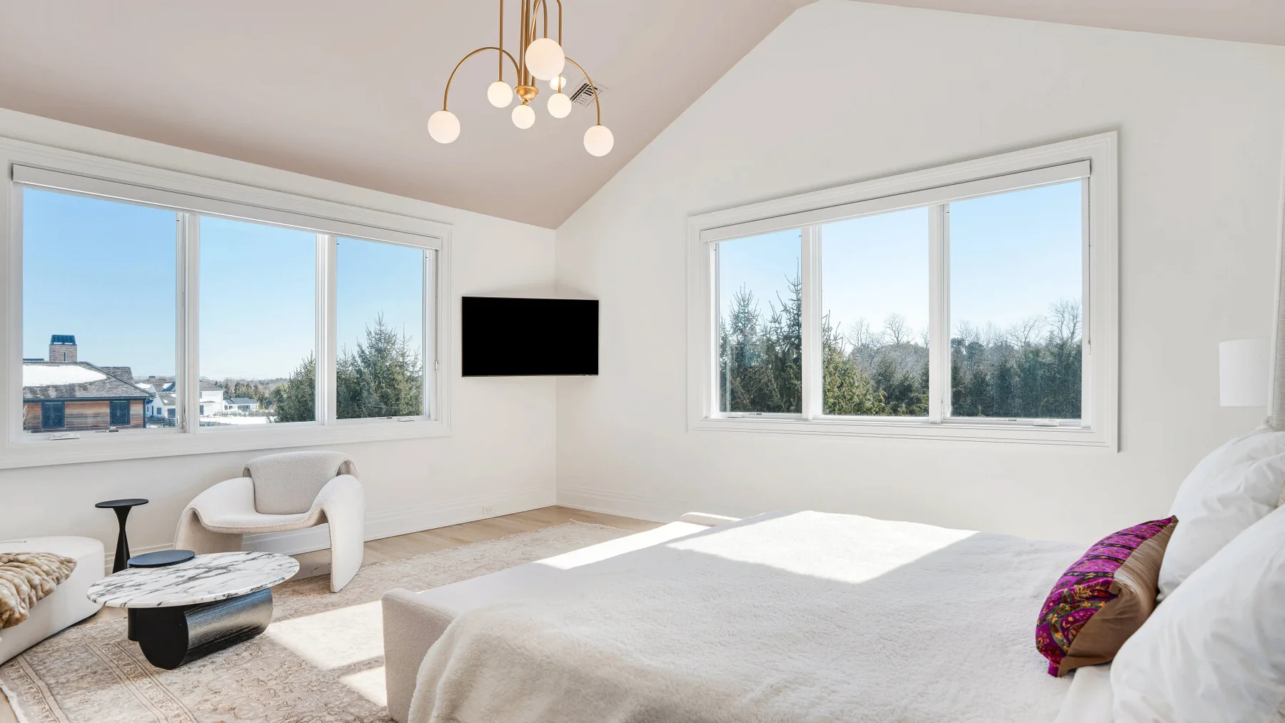 Un dormitorio luminoso con una cama, un televisor en la pared y ventanas grandes.
