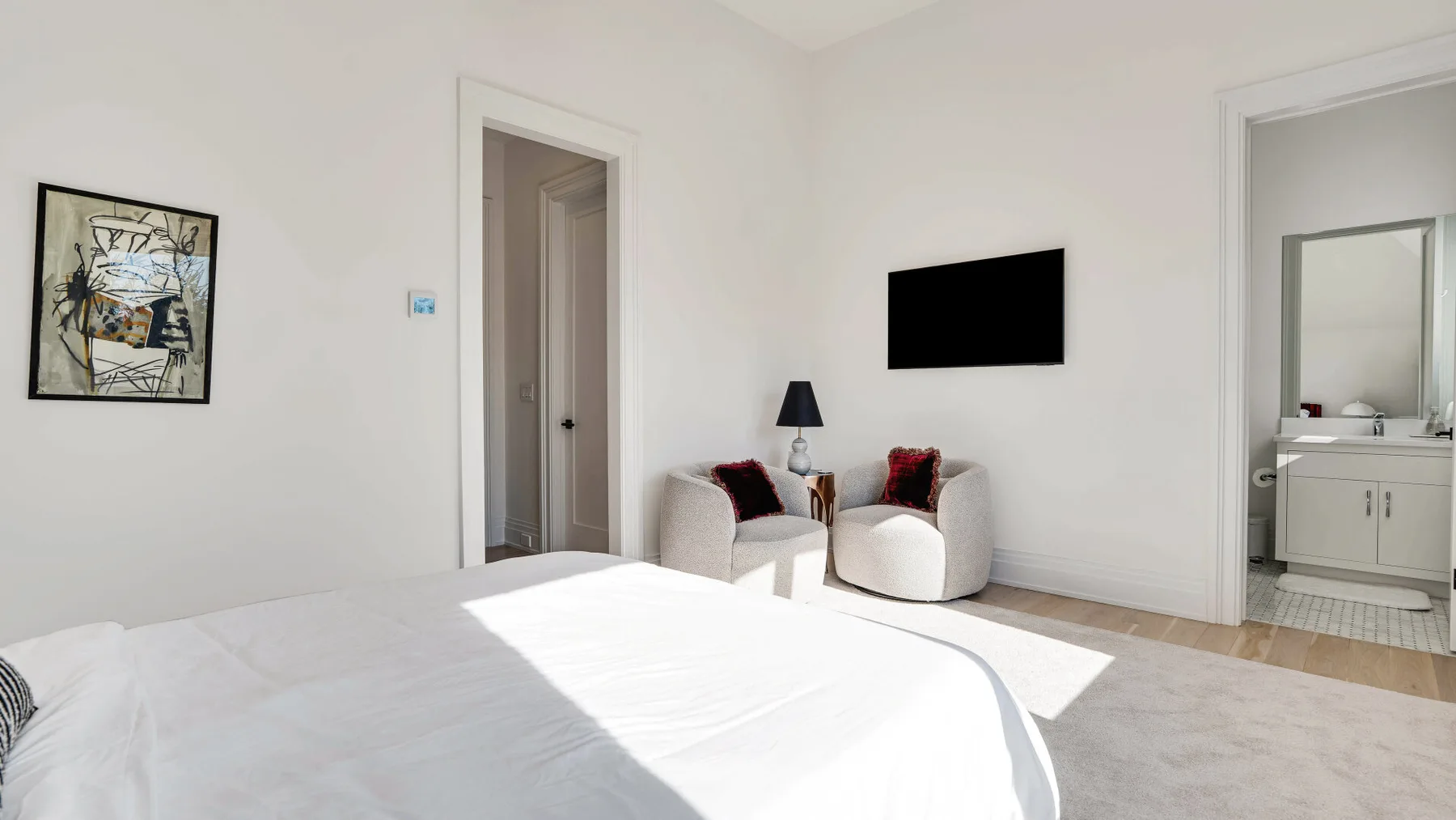 Un dormitorio luminoso con una cama blanca, dos sillones y un televisor en la pared.