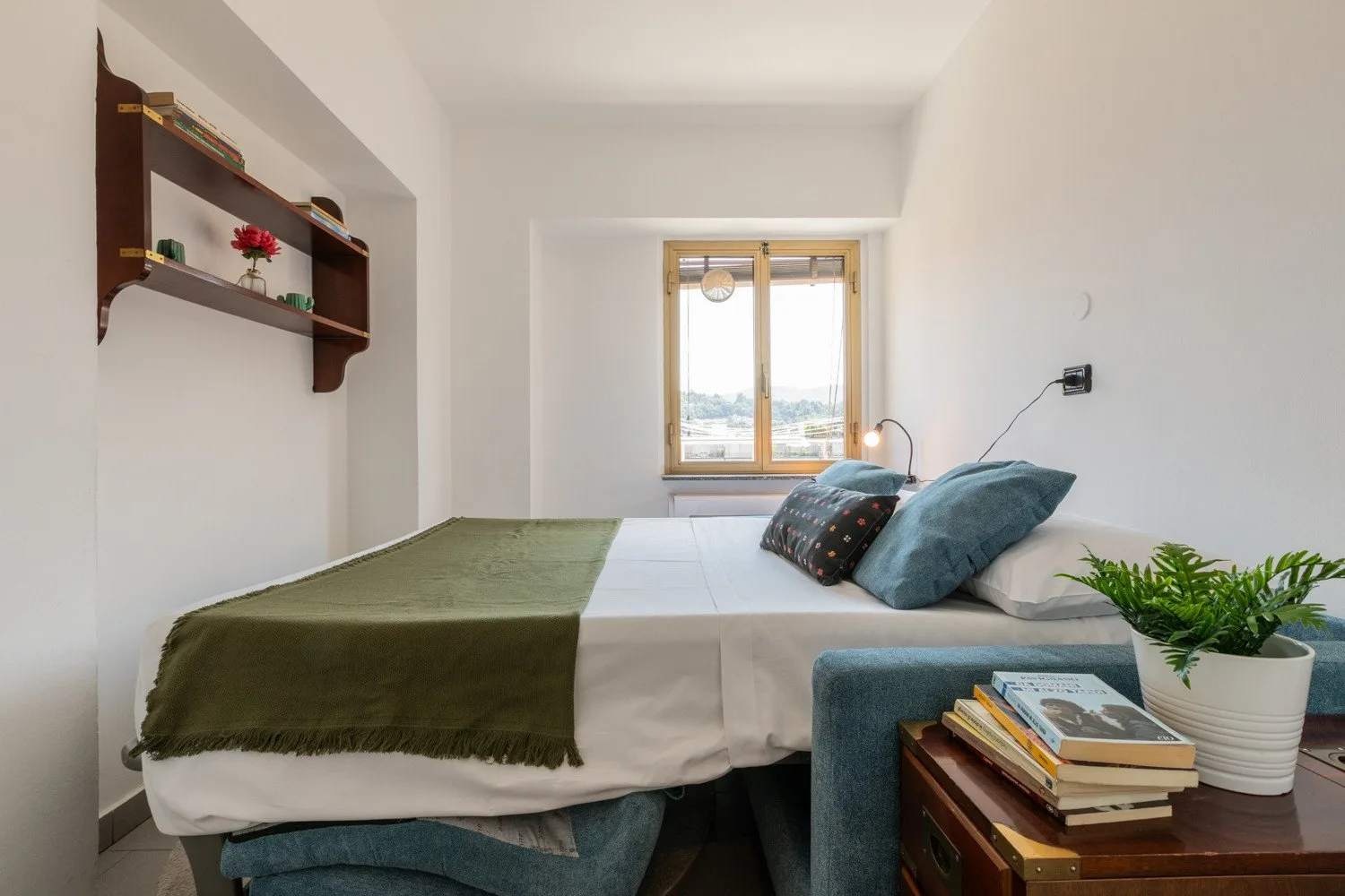 Un dormitorio con una cama, cojines verde azulado y una ventana con vista al exterior.