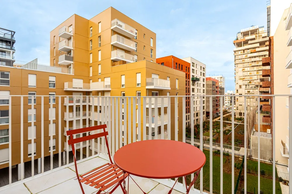 A balcony with a red table and chair overlooks nearby apartment buildings.