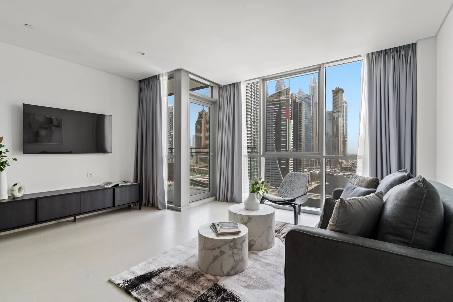 A bright living room with seating beside floor-to-ceiling windows overlooking city buildings.