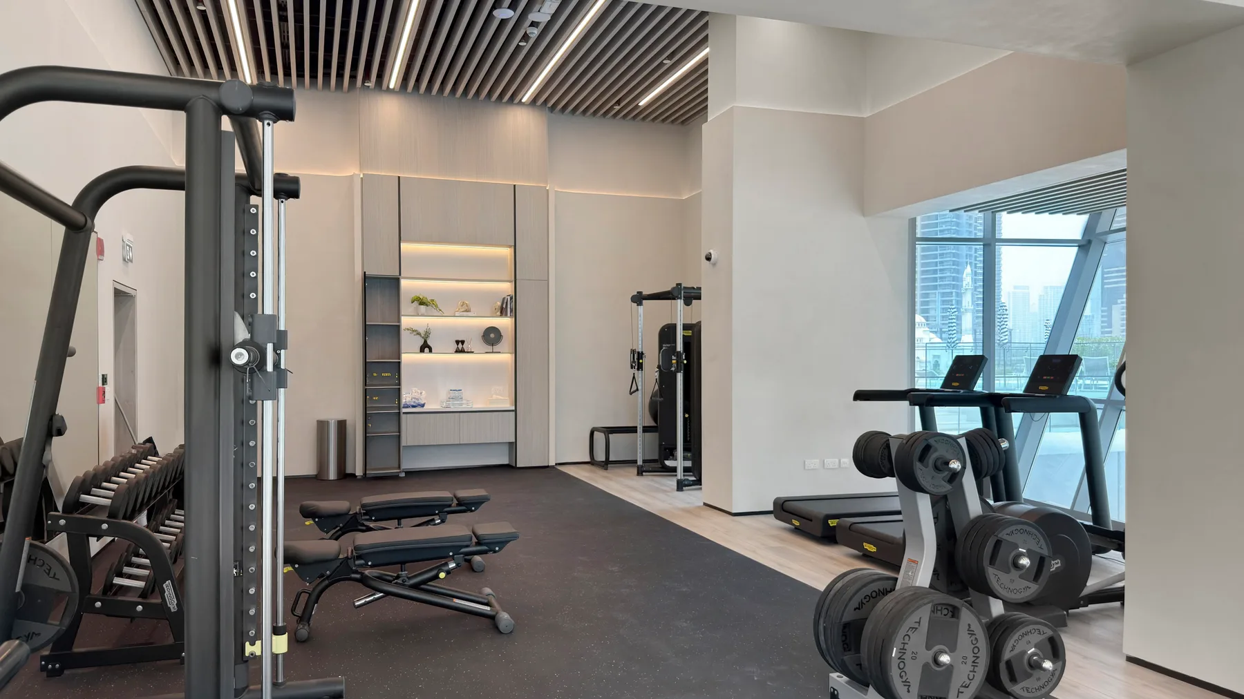 A modern gym with exercise machines, weights, and windows overlooking nearby buildings.