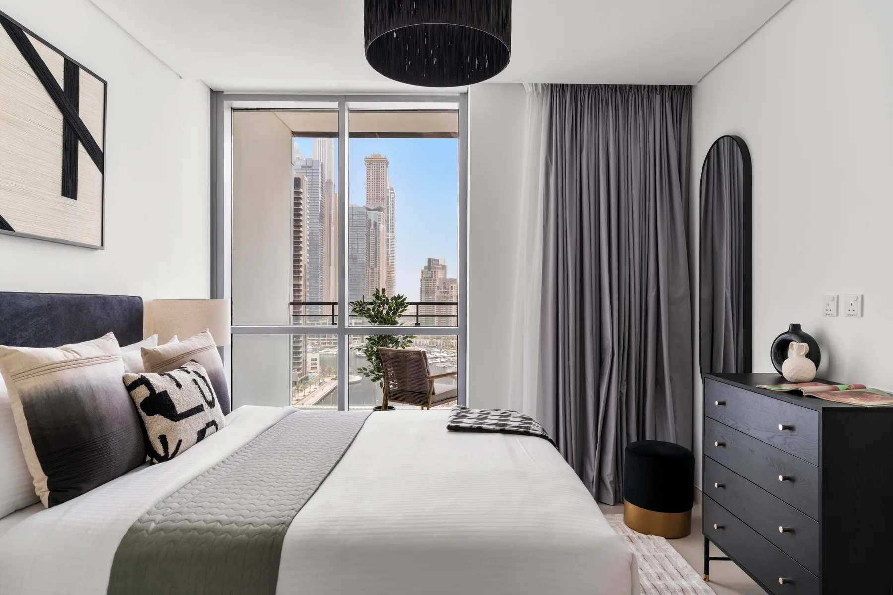 A bedroom with a neatly made bed beside a large window overlooking city buildings.