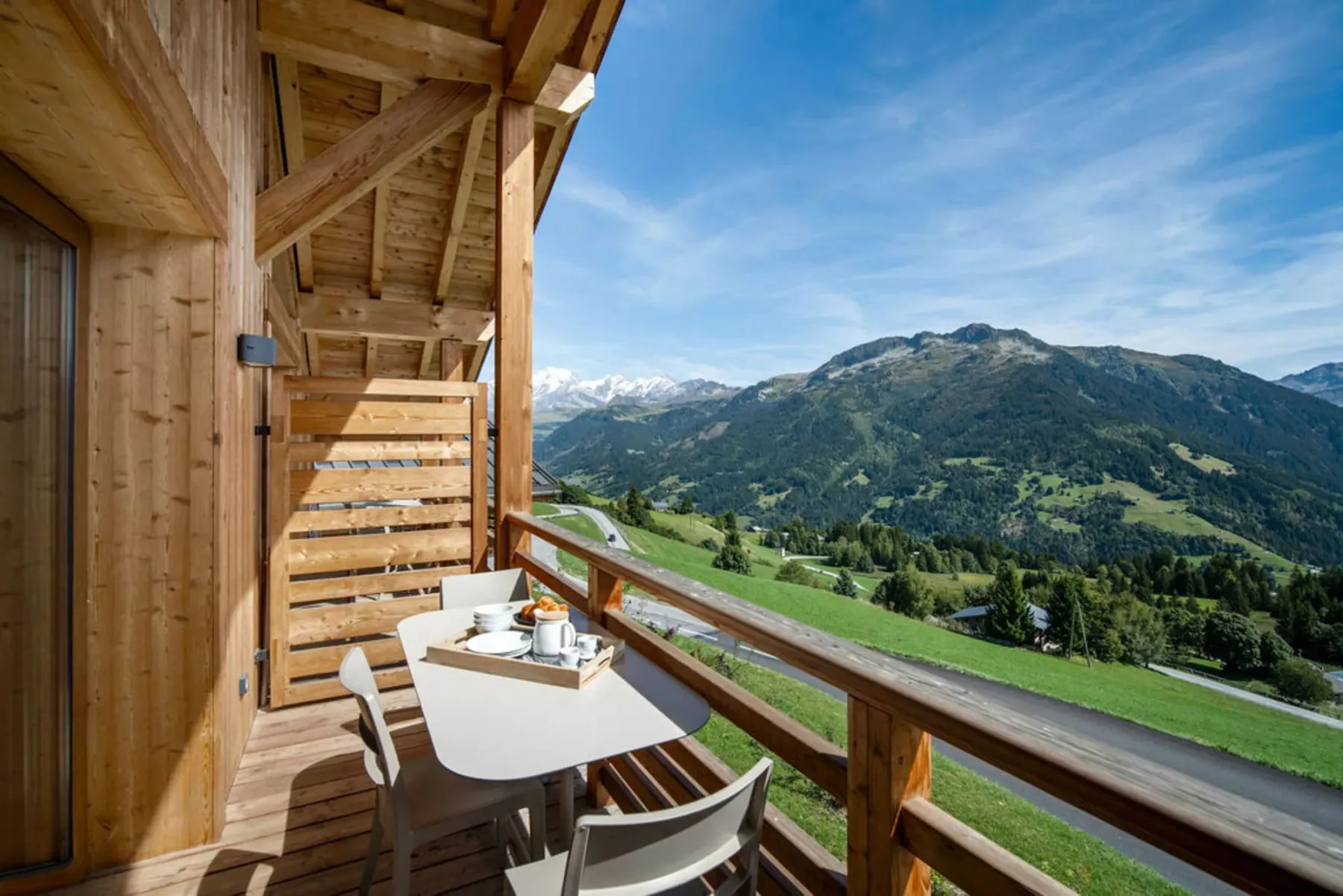 A wooden balcony with a small table overlooks a green mountain valley.