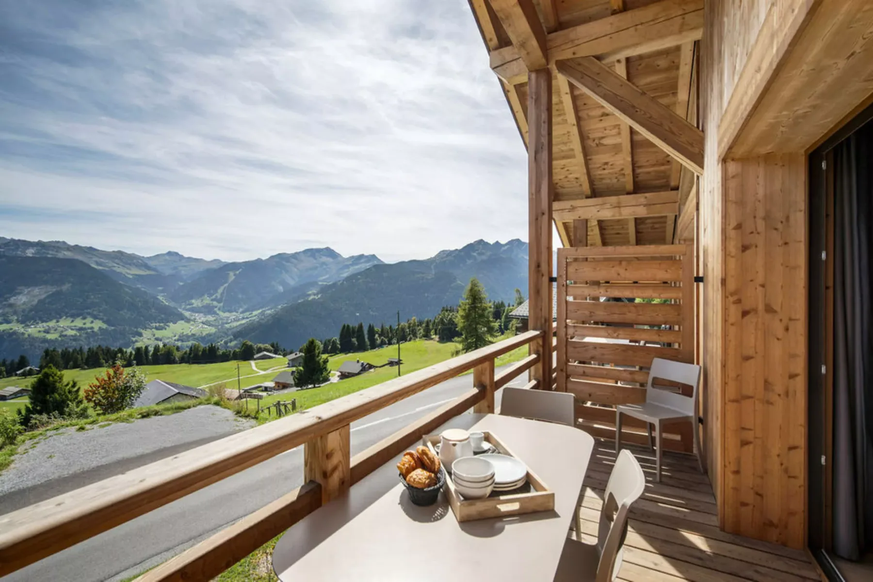 A wooden balcony with a dining table overlooks a green mountain valley.